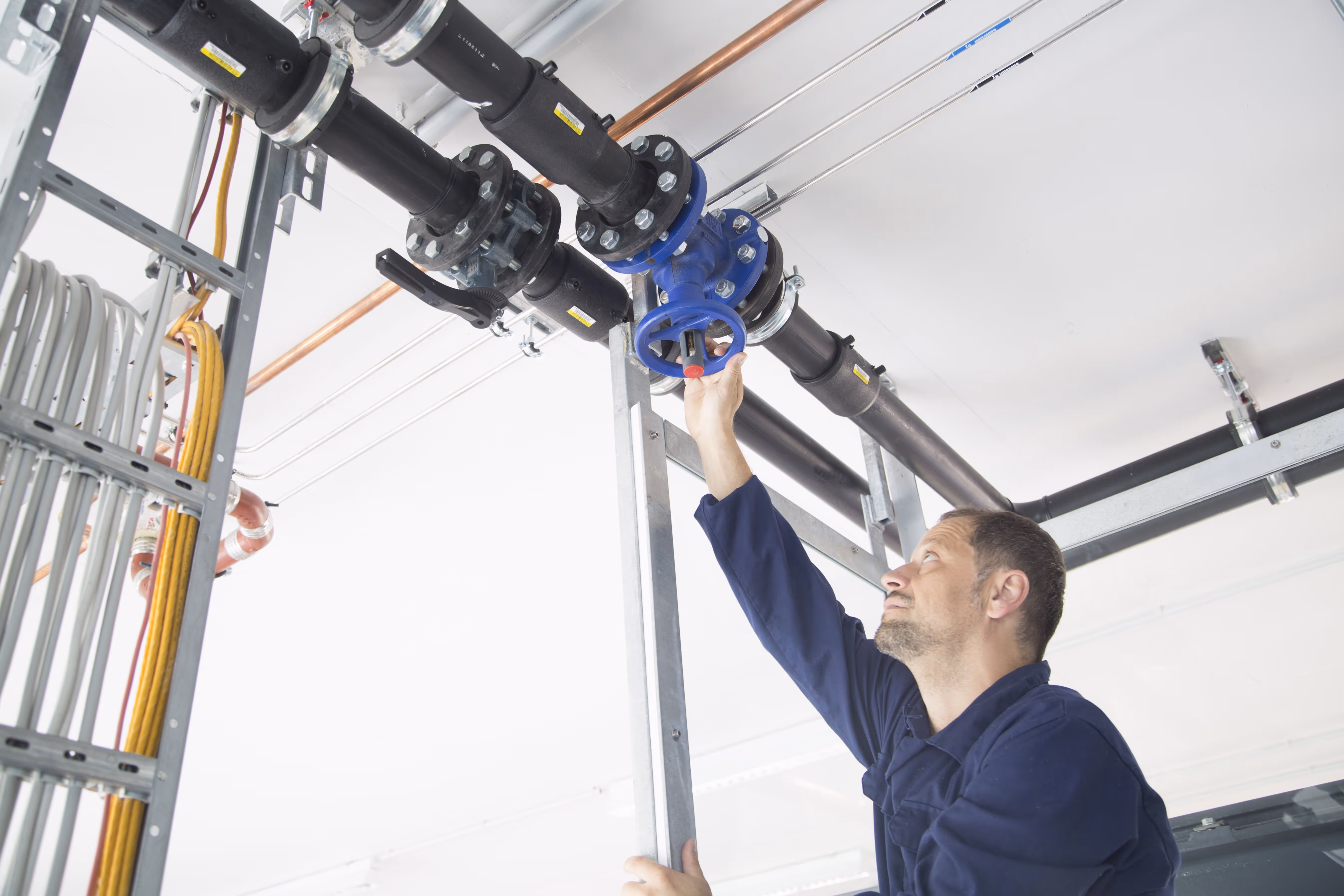 Maintenance technician adjusting a blue industrial valve on an overhead black pipe system, standing on a ladder inside a commercial or mechanical room.