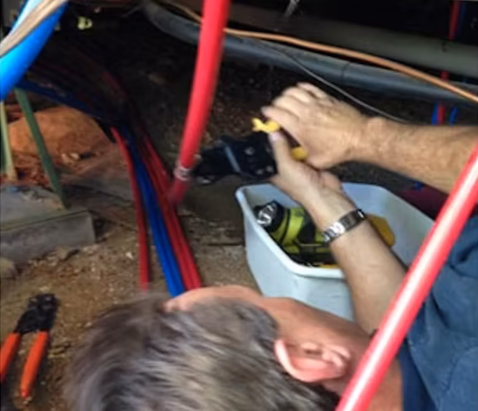 Plumber working in a crawl space using a crimping tool to install red PEX tubing, with blue PEX lines nearby and a white container holding tools and fittings.