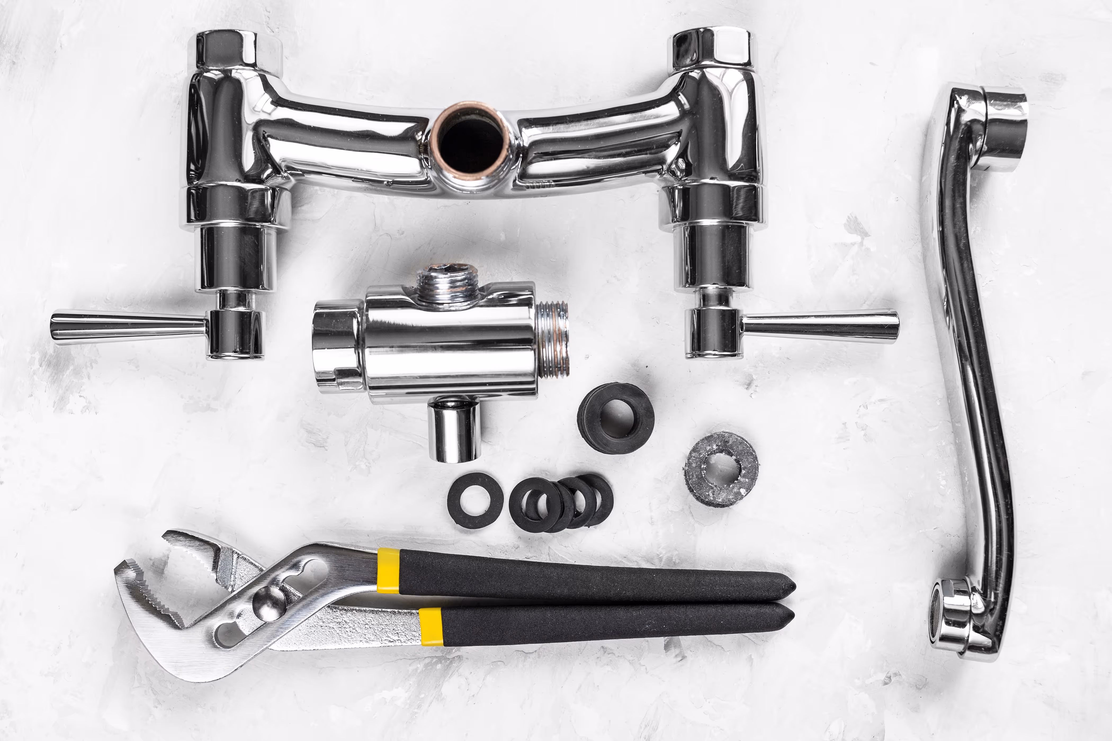 Plumbing toolset and chrome faucet components arranged on a white surface, including washers, an adjustable plier wrench, and a spout with handles.