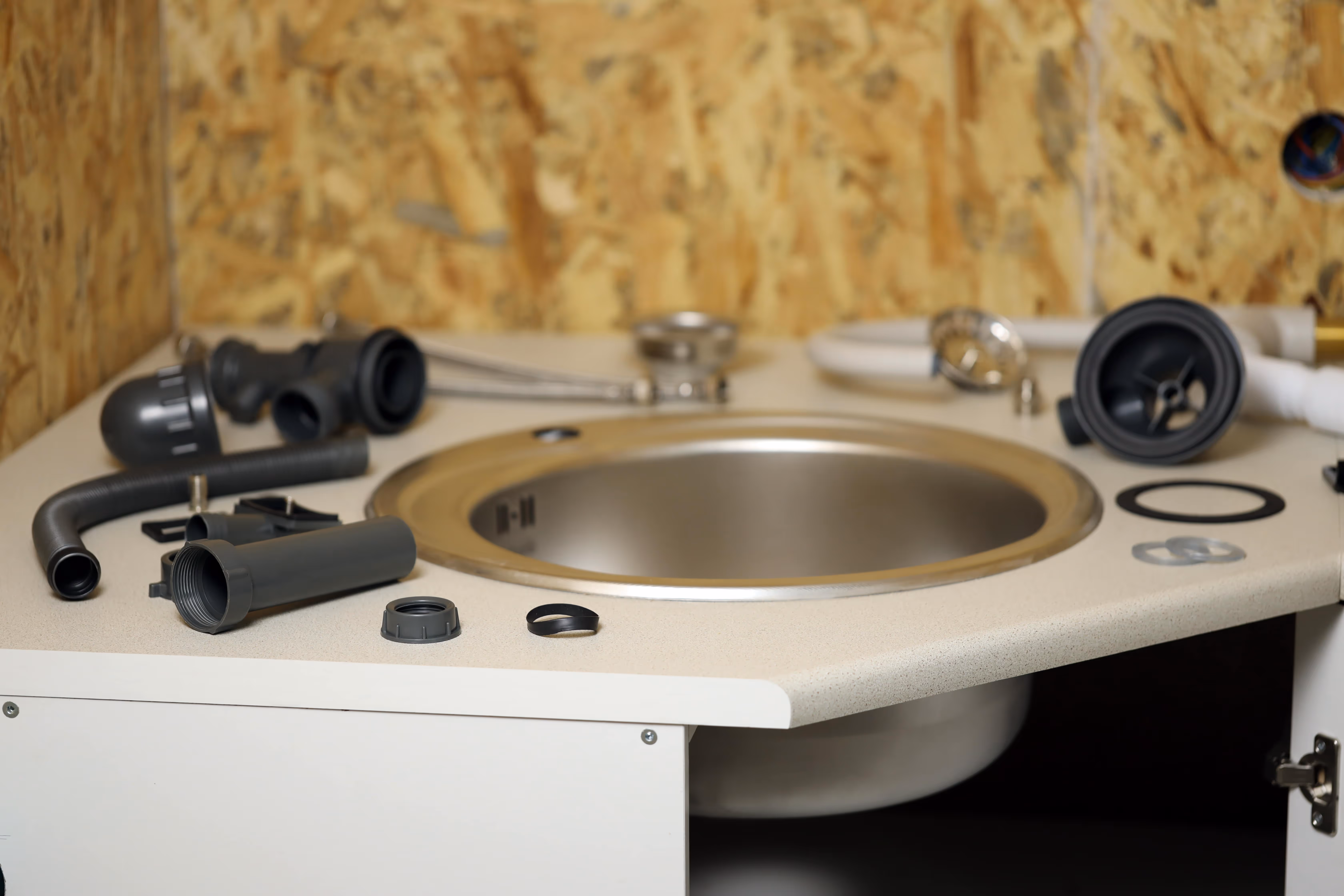 Uninstalled kitchen sink surrounded by loose plumbing components, including drain pipes, rubber gaskets, and fittings, on a countertop with an exposed plywood backsplash.