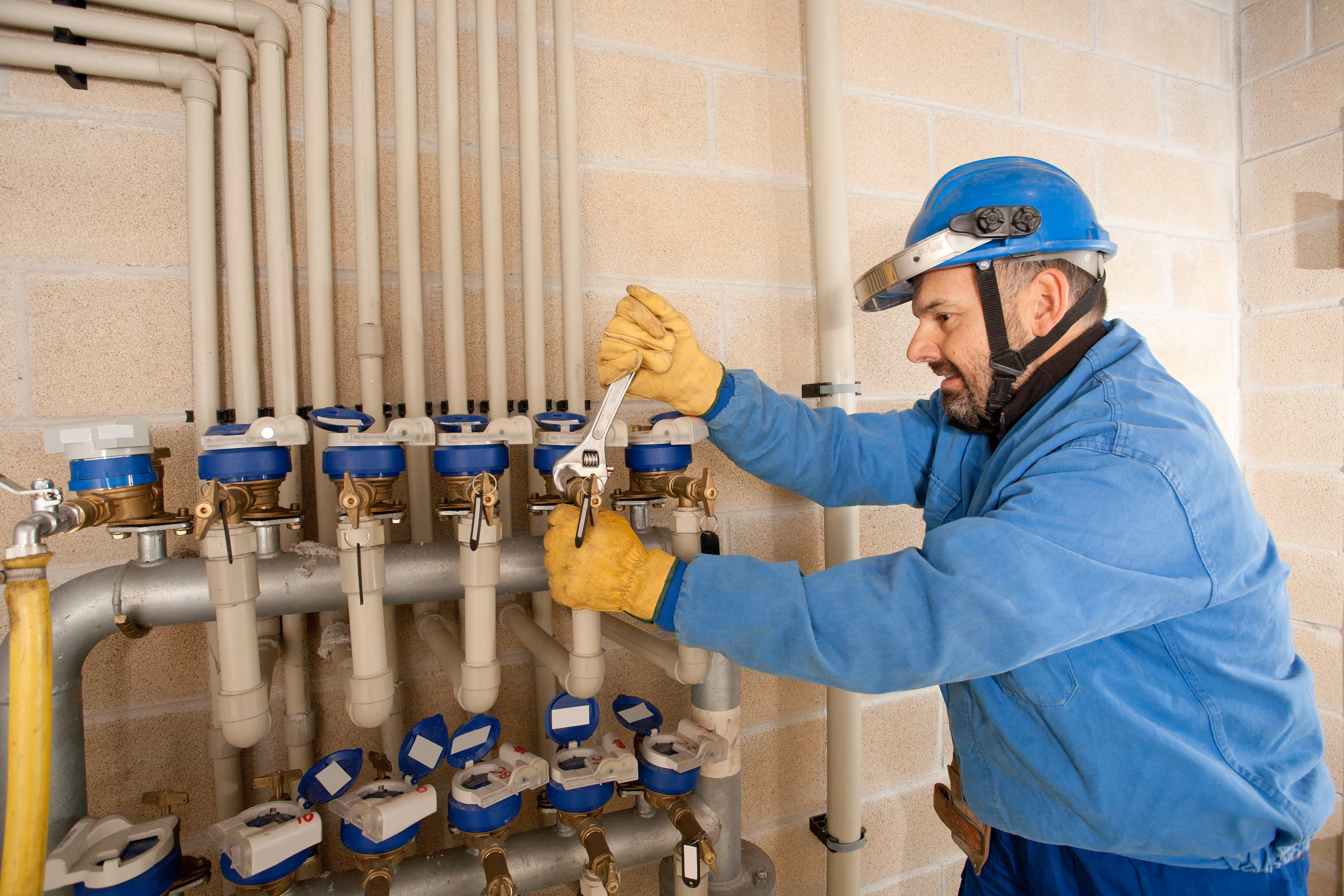 Plumber in protective gear adjusting a manifold of water meters and PVC pipes using a wrench, inside a utility room with exposed brick walls.