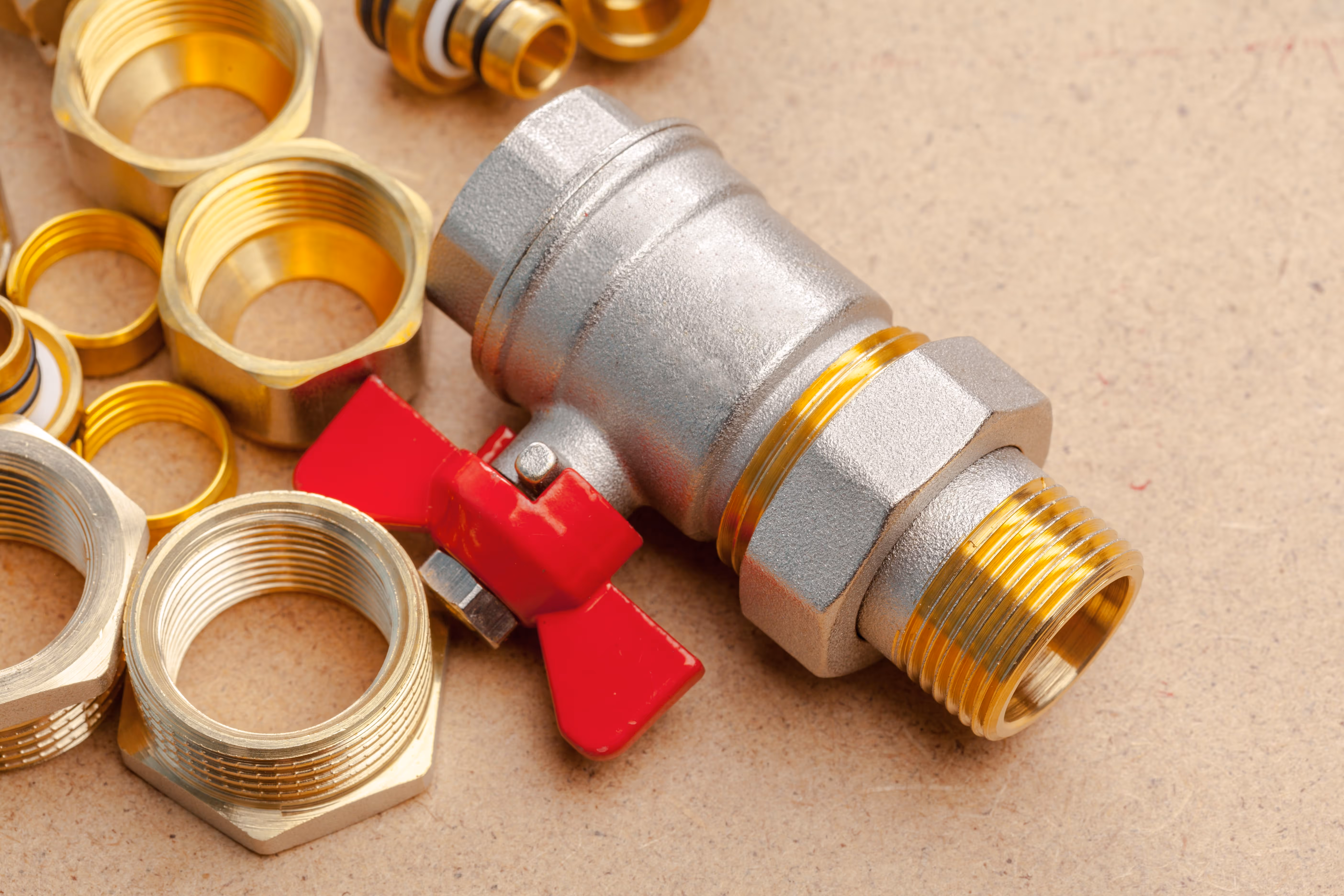 Close-up of a metallic ball valve with a red butterfly handle surrounded by brass plumbing fittings on a workbench surface.