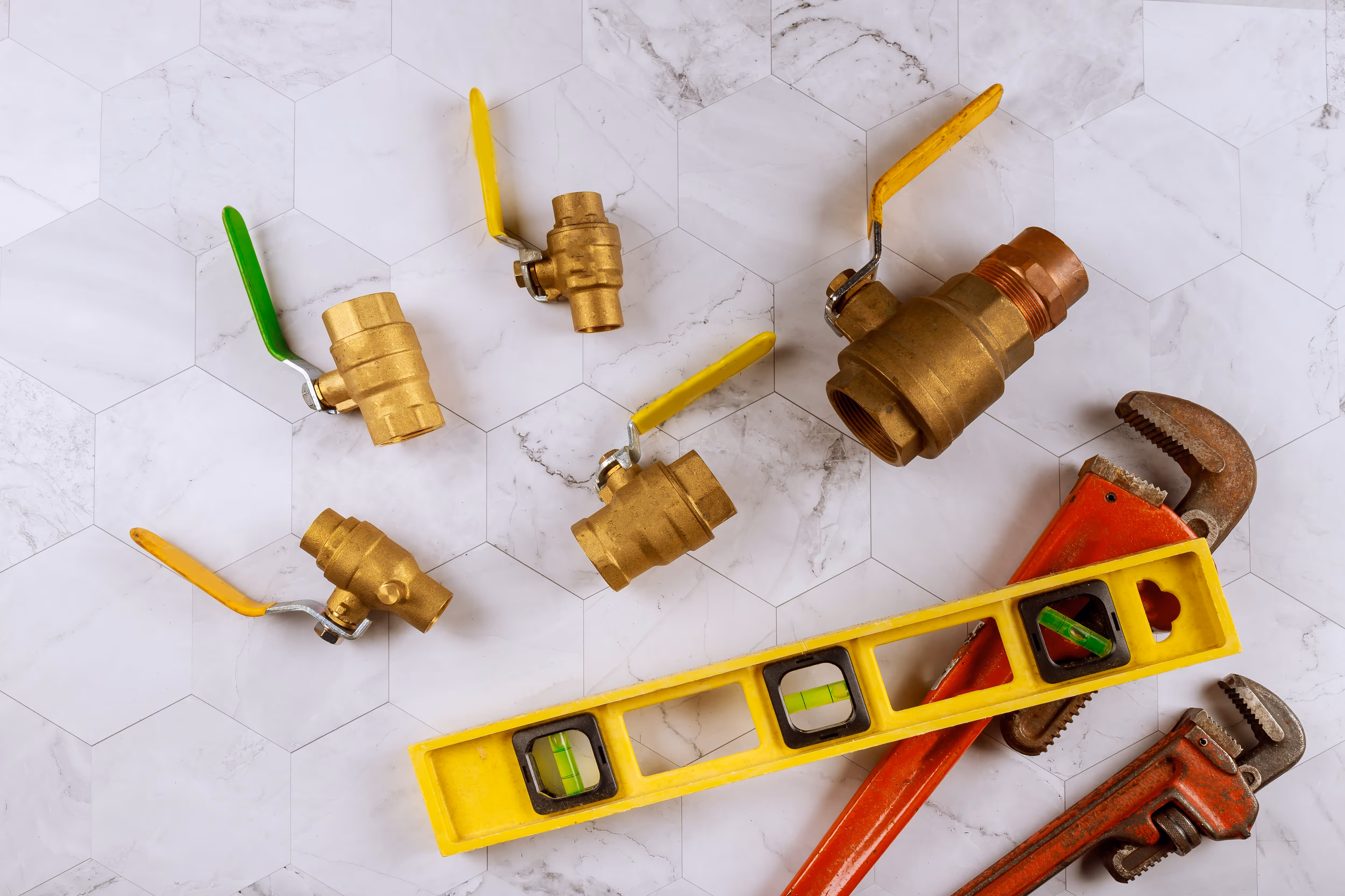 Collection of brass ball valves with yellow and green handles arranged on a tiled surface, alongside a yellow spirit level and two red pipe wrenches.