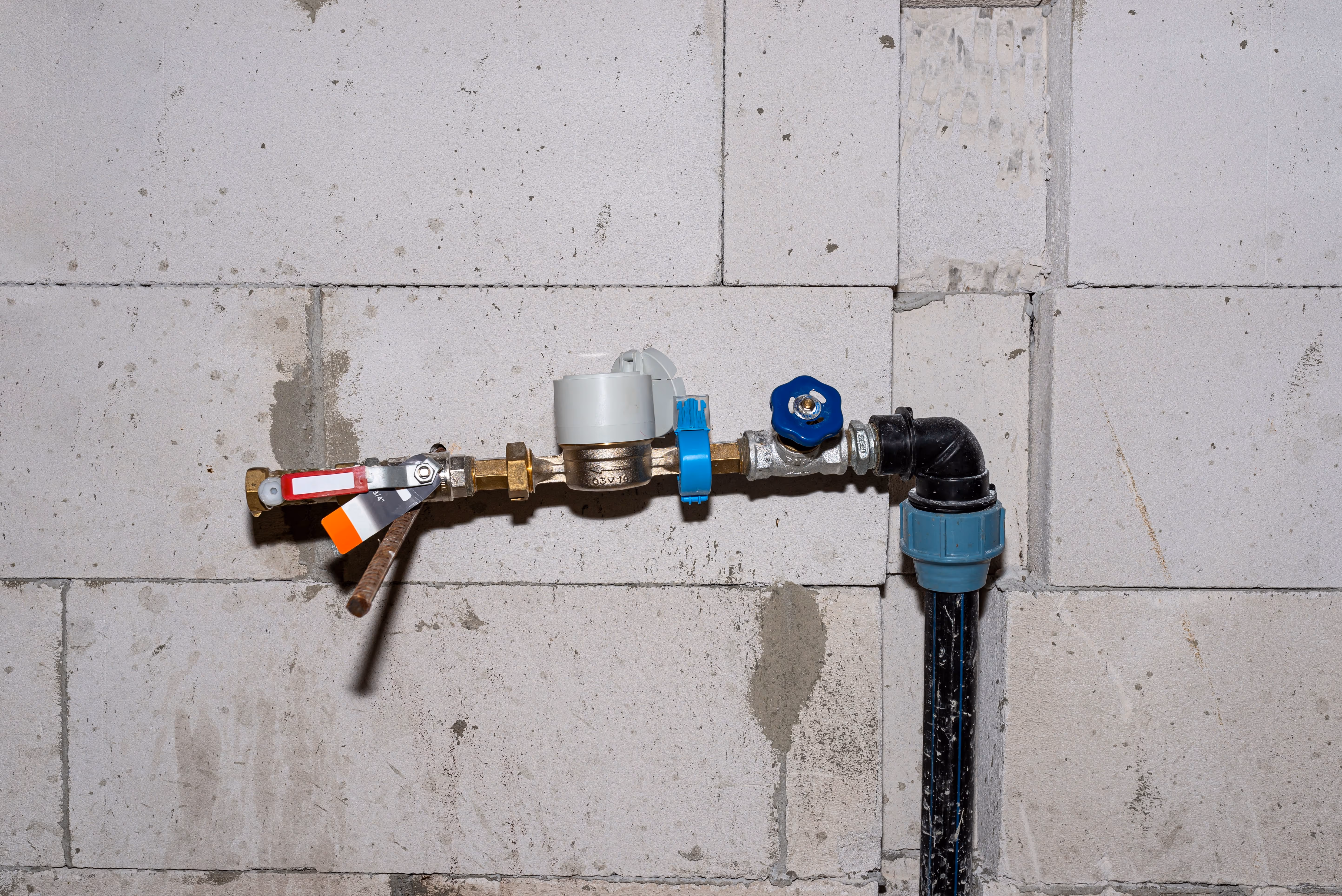 Close-up of a domestic water shutoff valve and meter assembly mounted on a concrete wall, with brass fittings, a red-handled ball valve, blue plastic connector, and black polyethylene piping.