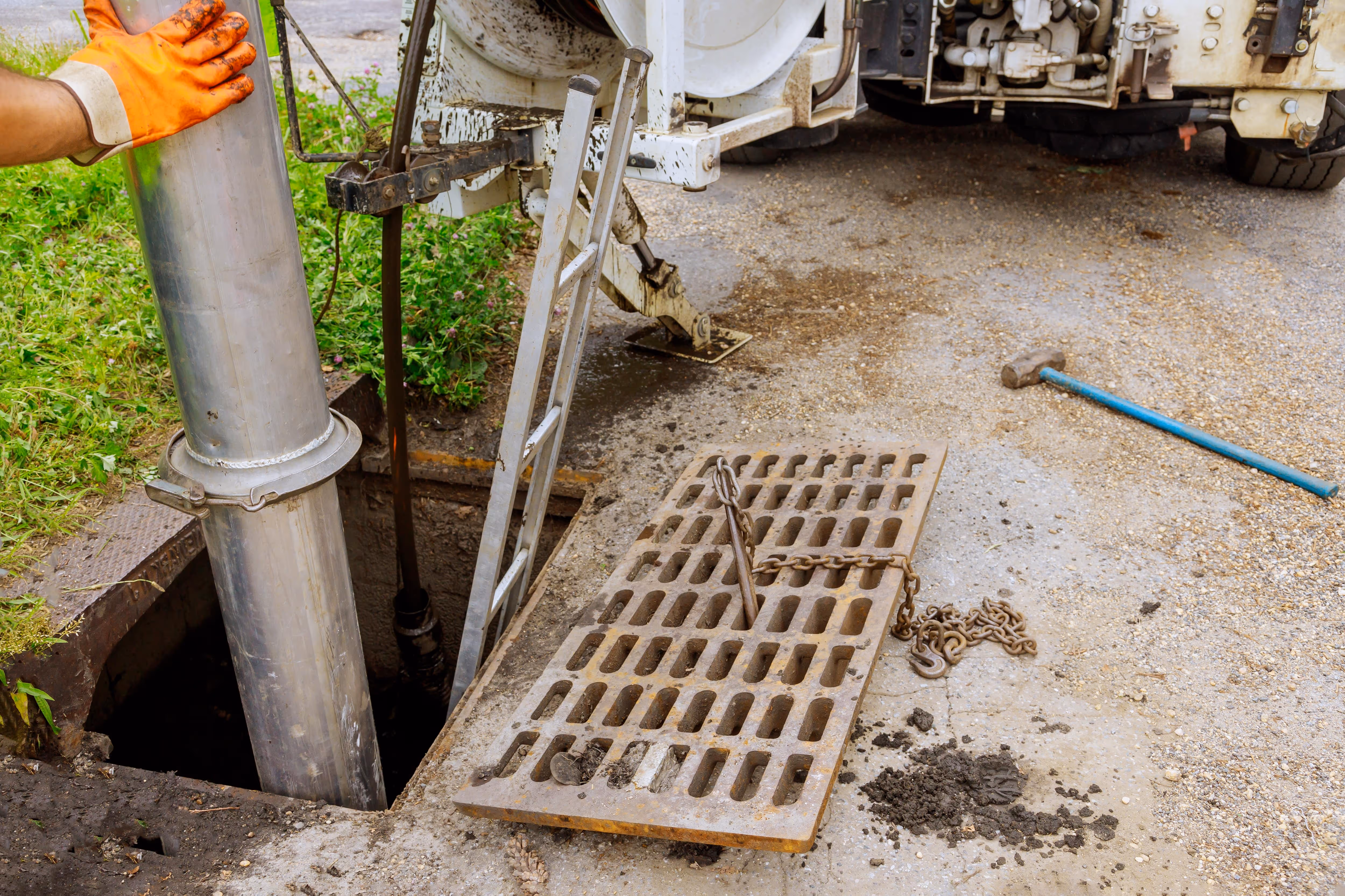 A utility technician in safety gear using a sewer‑cleaning vacuum truck to access a sewer manhole and flush blockages with high-pressure piping.