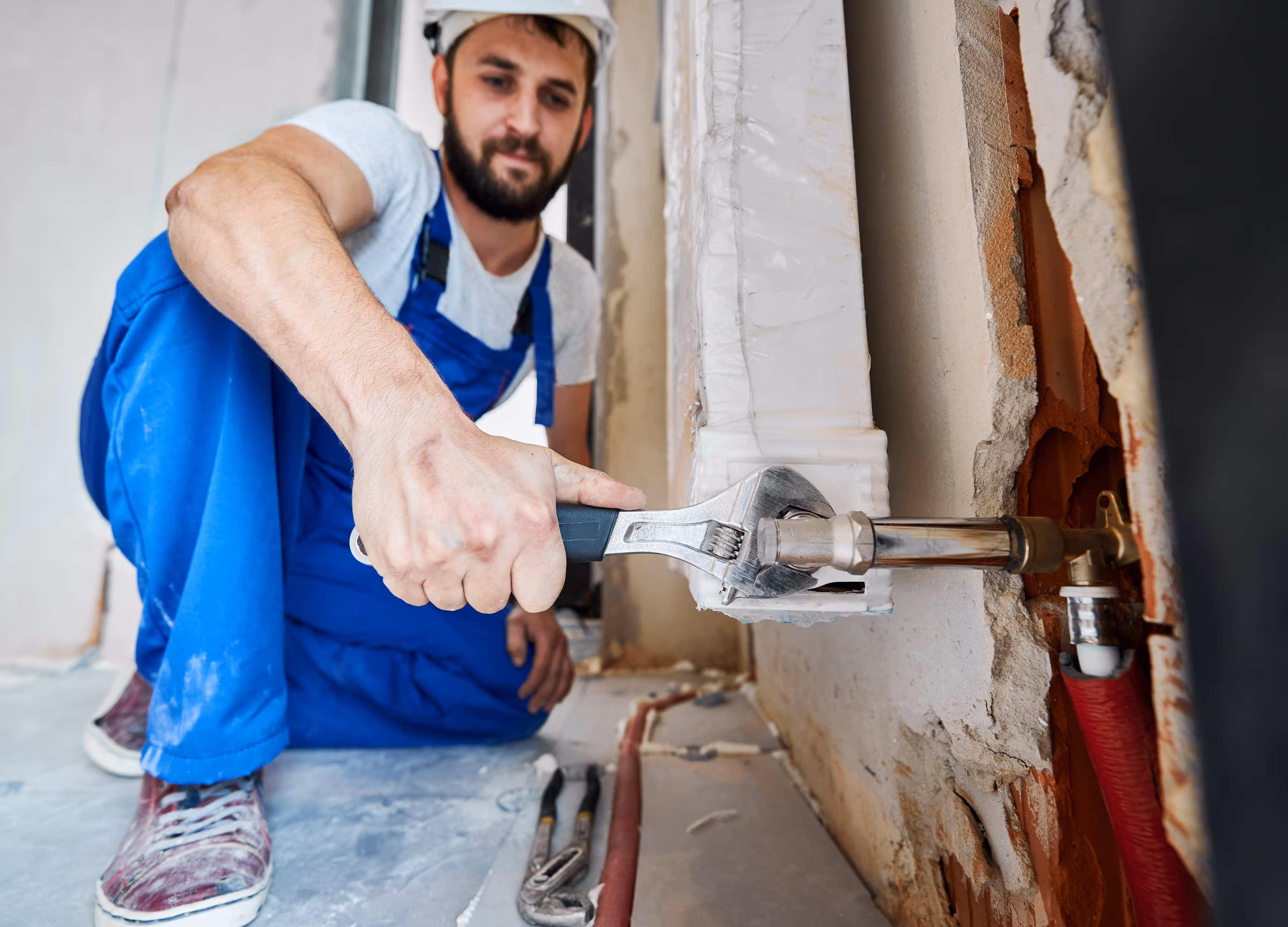 A plumber installing a heating radiator in an apartment, using tools to secure the unit to the wall with exposed piping visible.