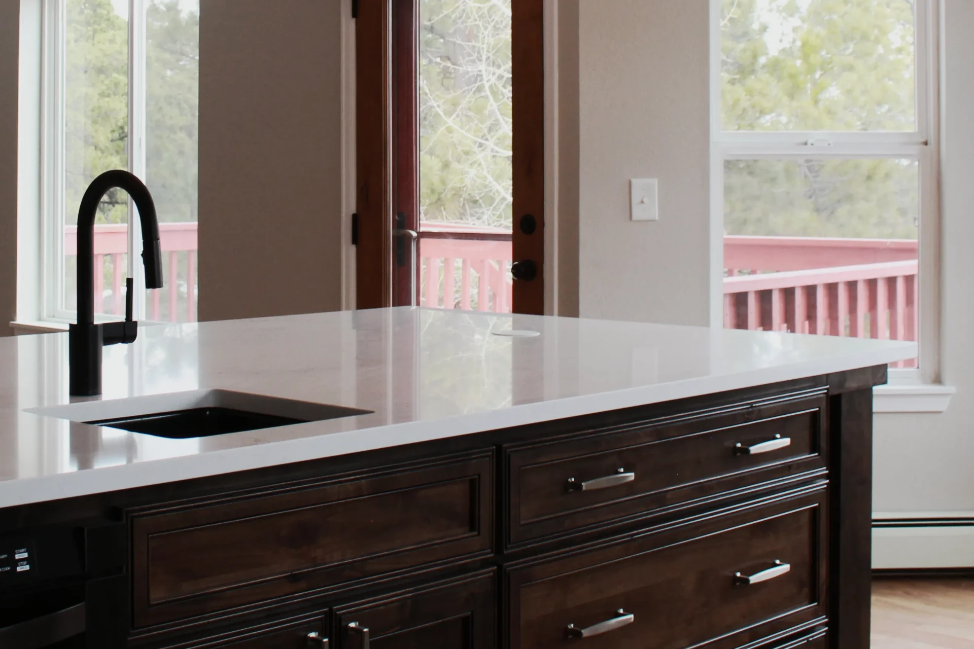 Modern kitchen island with dark wood drawers, white countertop, and black faucet, near windows and a glass door.