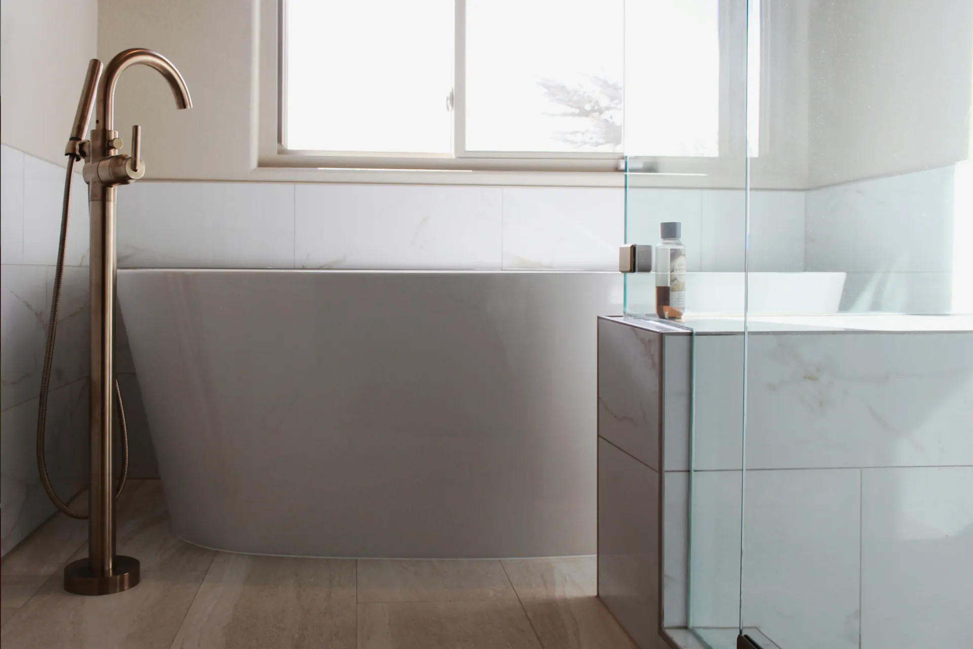 Modern freestanding bathtub with gold-tone floor-mounted faucet and handheld shower next to a glass shower enclosure in a bright bathroom.