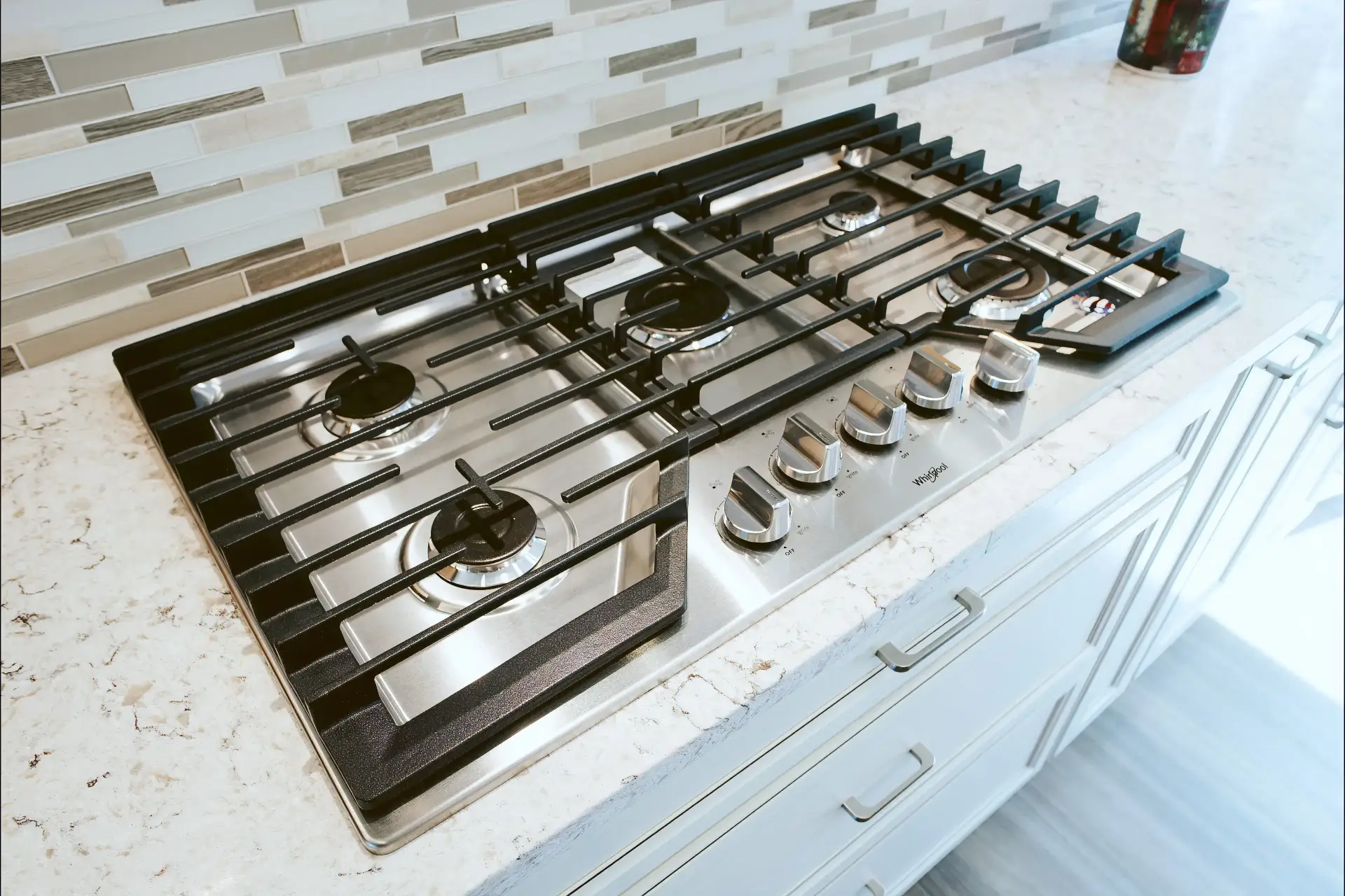 Stainless steel Whirlpool gas cooktop with five burners and black grates installed in a white granite countertop with beige and white tile backsplash.