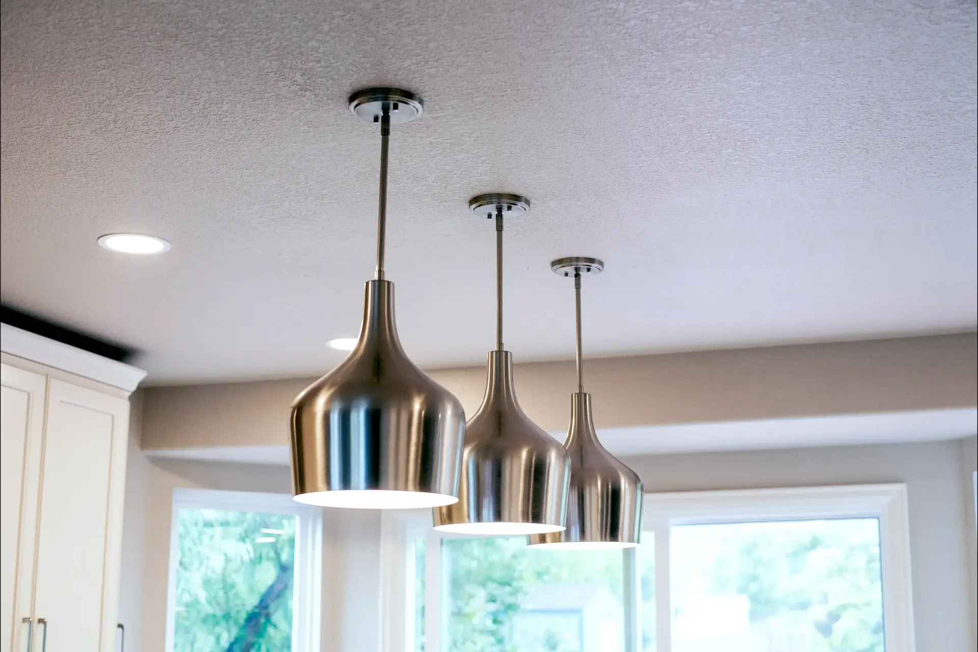 Three modern stainless steel pendant lights hanging from a kitchen ceiling above bright windows.