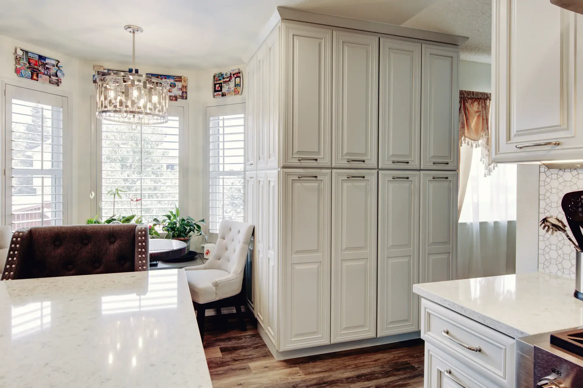 Bright kitchen corner with tall white cabinetry, white marble countertops, a crystal chandelier, and upholstered chairs near windows with shutters.