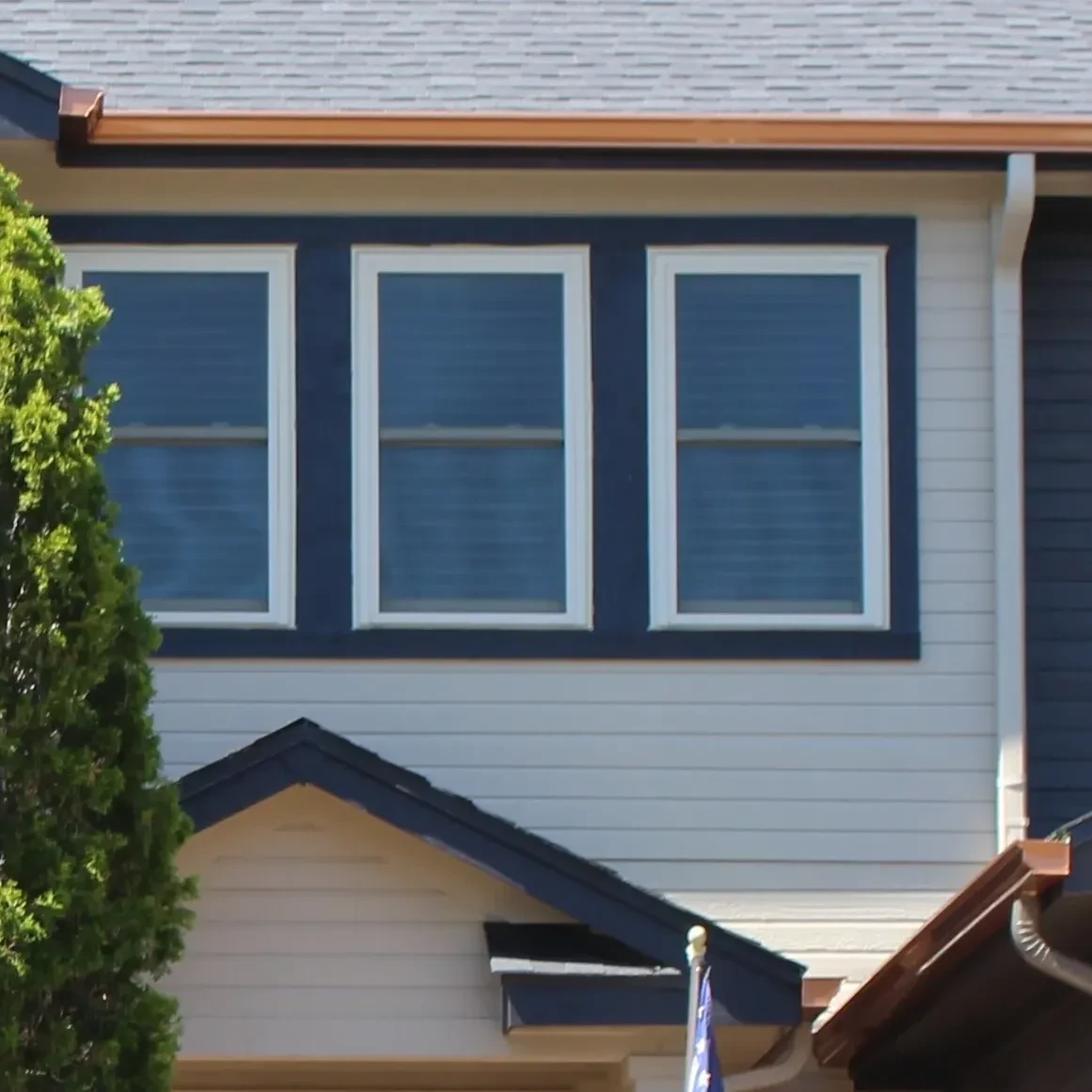 Upper facade of a house featuring three white-trimmed windows with blue shutters, beige siding, a small roof overhang, and part of a green tree on the left.