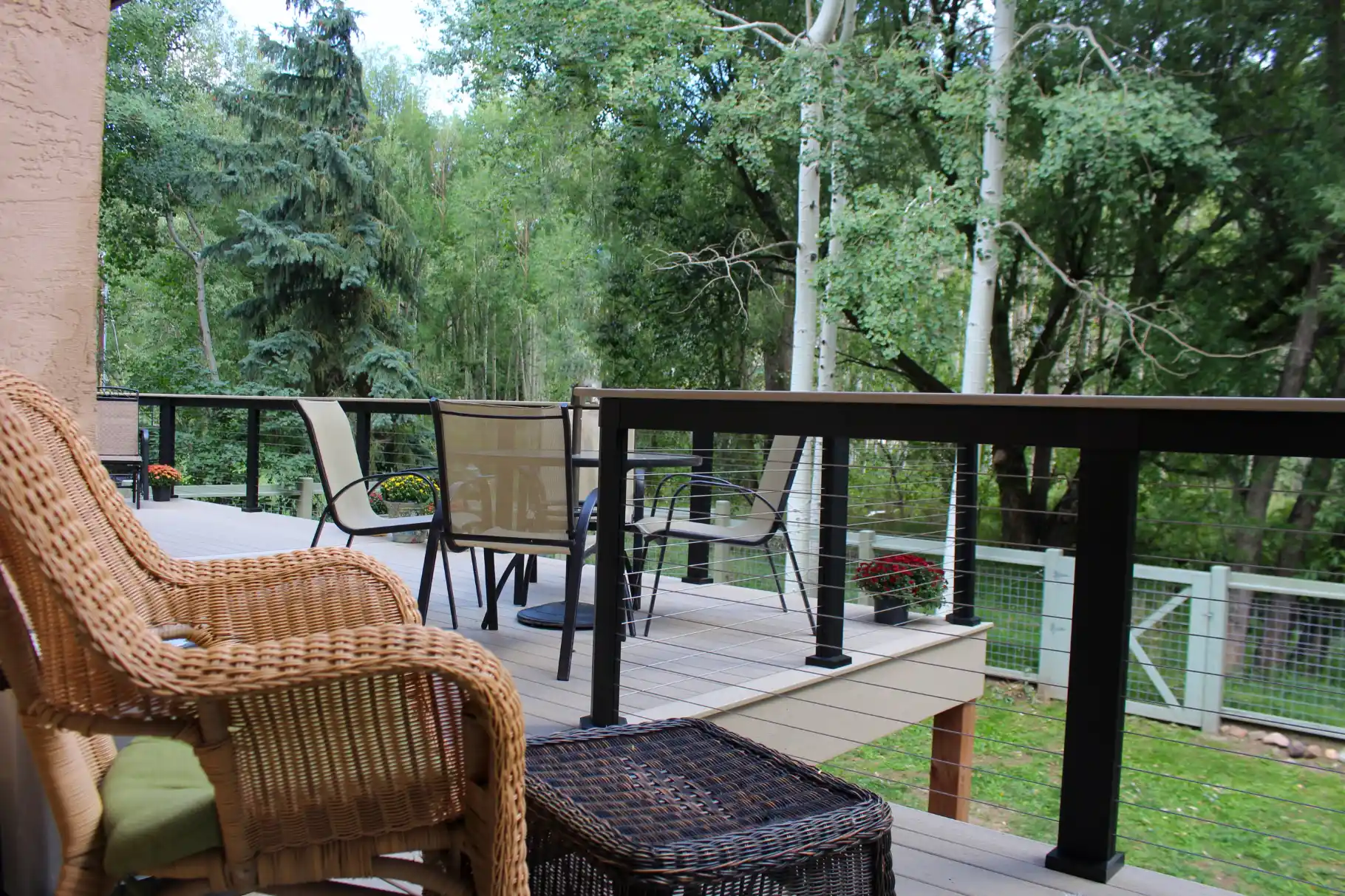 Outdoor deck with wicker chair, table, and metal railing overlooking a green backyard with trees.