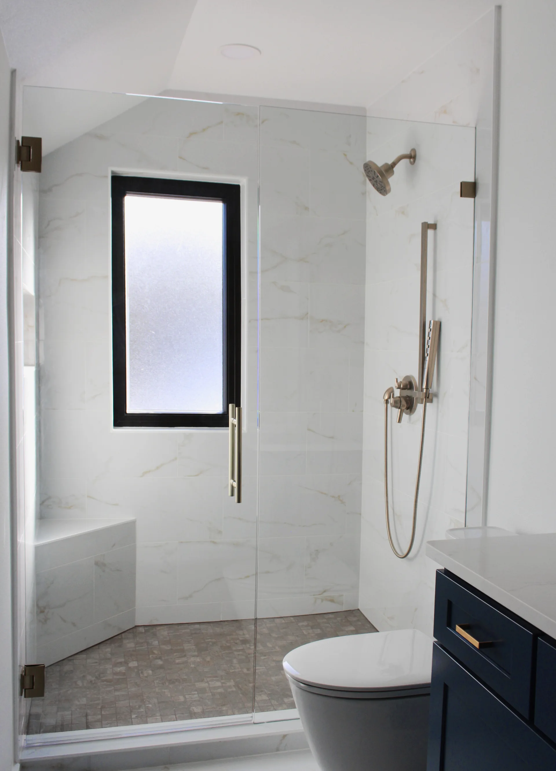 Modern bathroom with glass shower enclosure, marble-patterned tiles, a bench, and brushed metal fixtures.