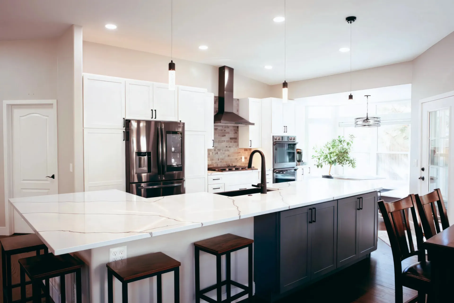 Modern kitchen with white and navy cabinets, marble countertop island with bar stools, black appliances, and large windows letting in natural light.