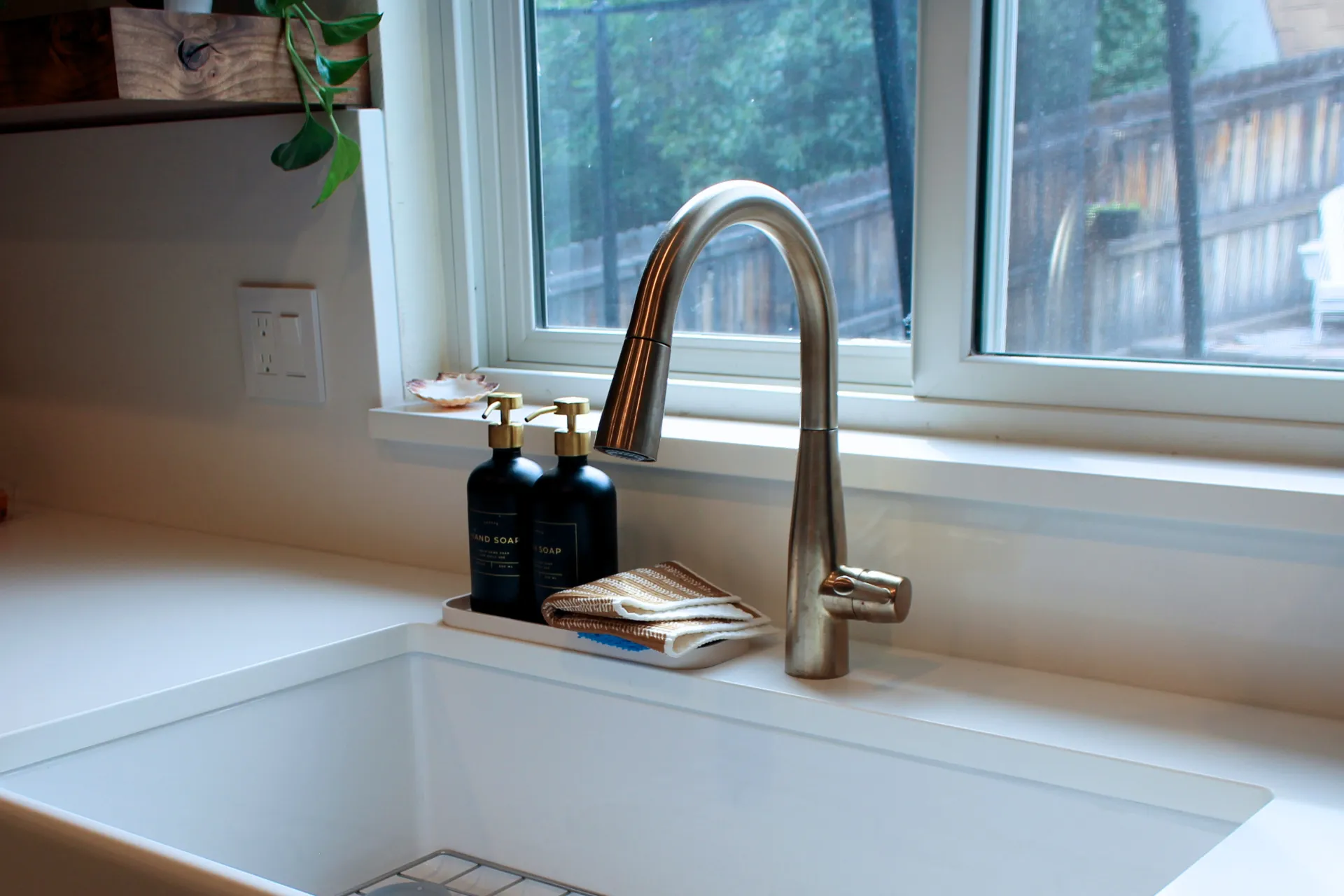 Modern kitchen sink with stainless steel faucet, two black soap dispensers, and a folded kitchen towel on the countertop under a window.