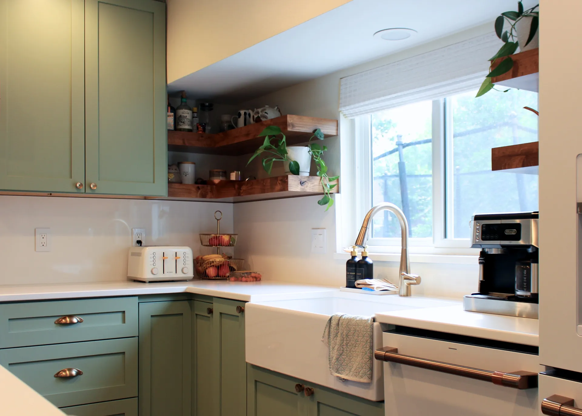 Modern kitchen corner with green cabinets, farmhouse sink under window, wooden shelves with plants, toaster, coffee maker, and fruit basket.