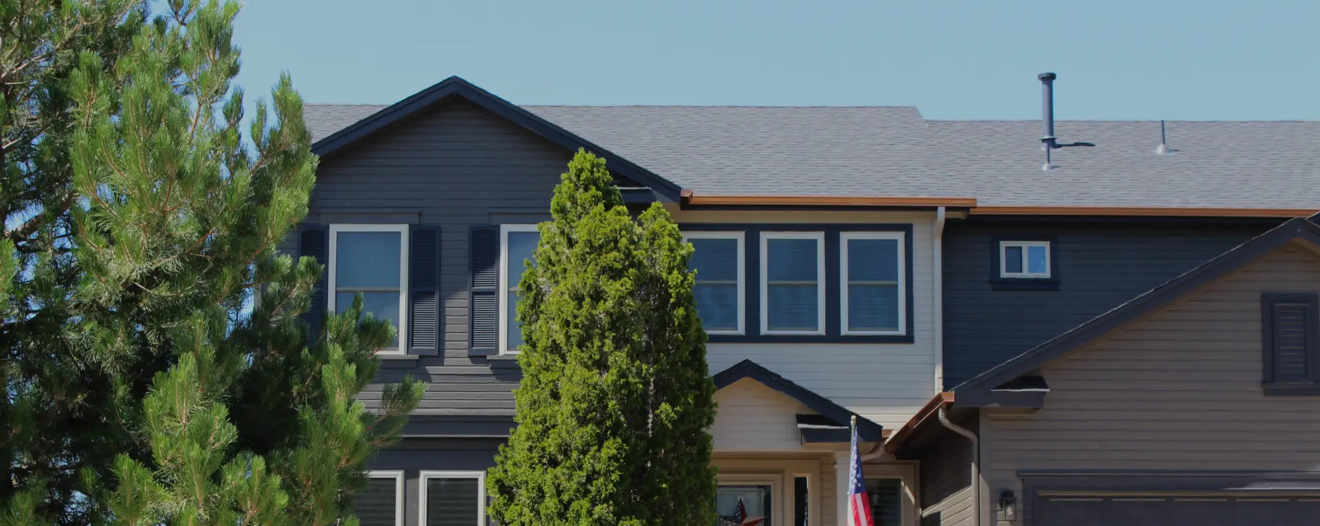 Two-story house with dark siding, multiple windows, copper gutters, and tall green pine trees in front.