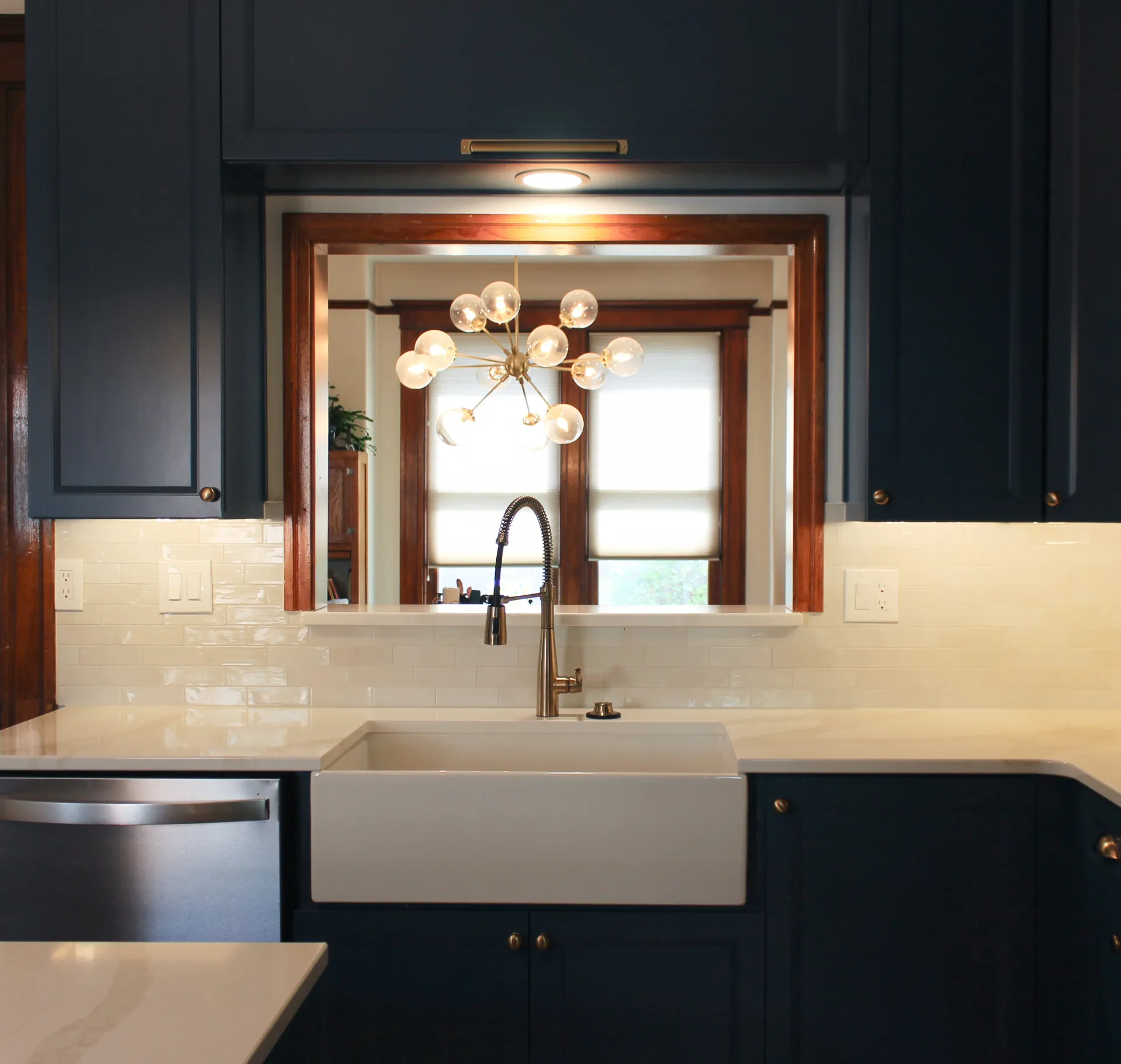 Modern kitchen sink area with white farmhouse sink, brushed metal faucet, dark blue cabinets, and view of a chandelier through a window opening.
