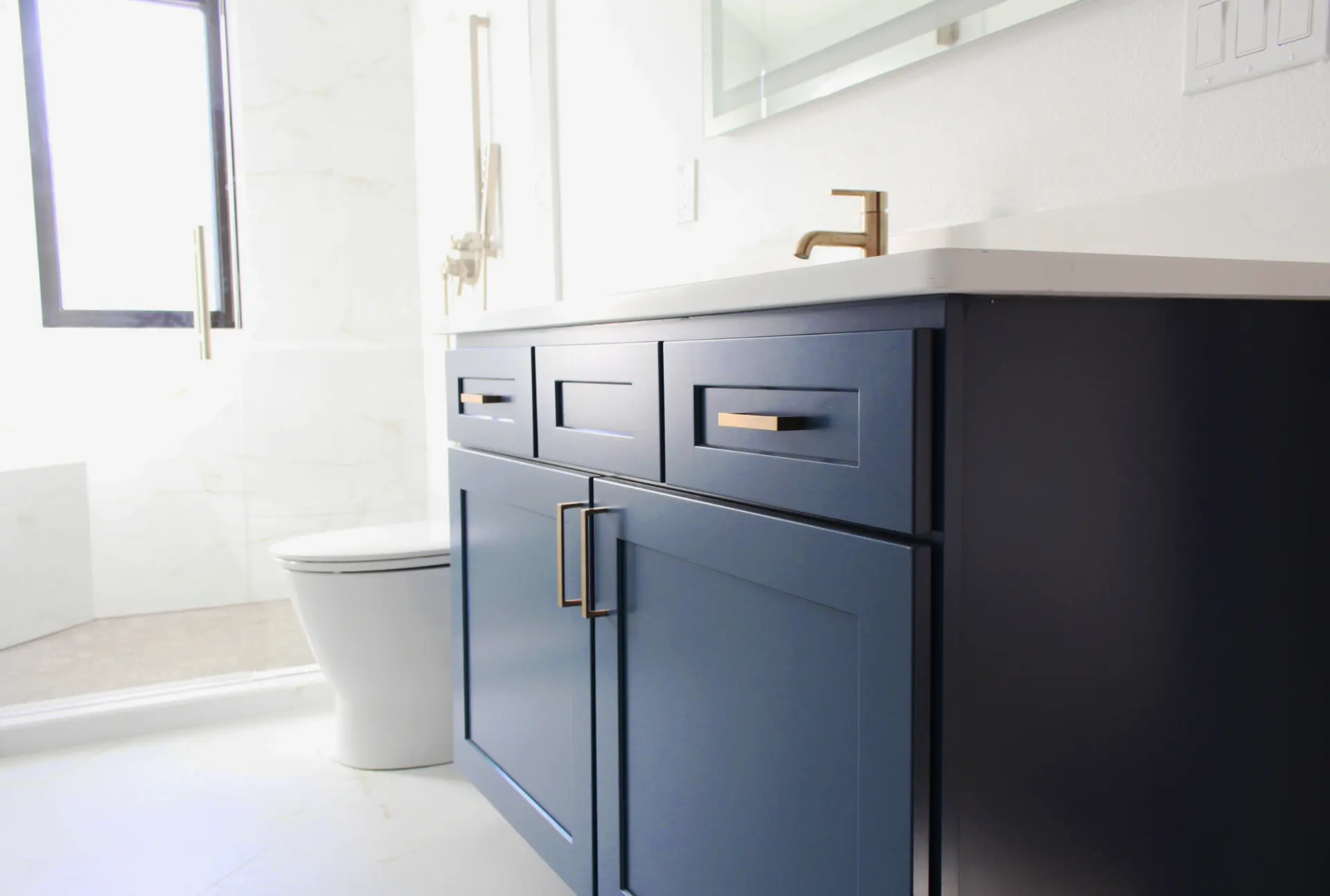 Bathroom with navy blue vanity featuring gold handles, white countertop, and a white toilet next to a glass shower door.