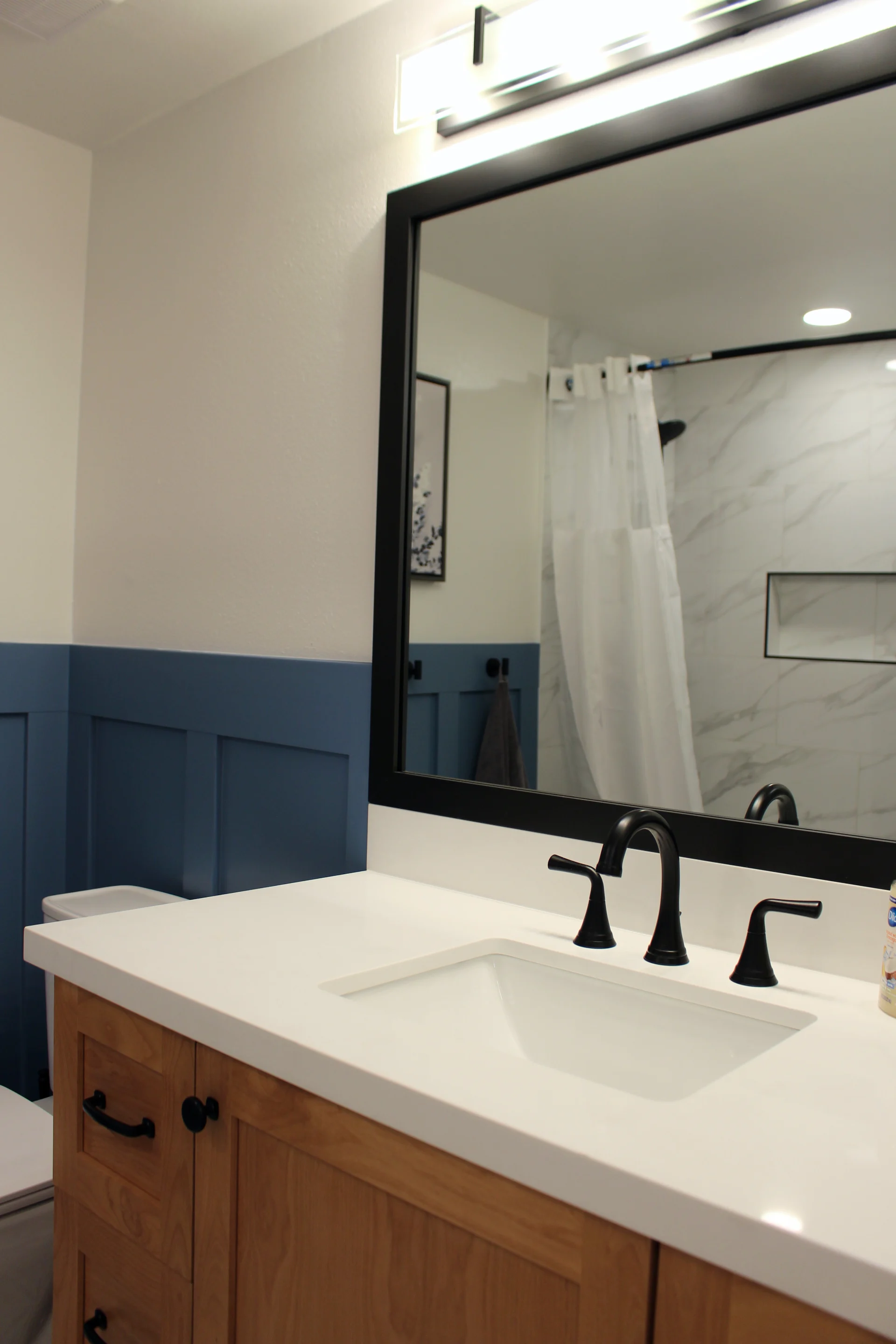 Bathroom vanity with white countertop, black faucet, wooden cabinet, and large black-framed mirror reflecting a shower with white curtain.