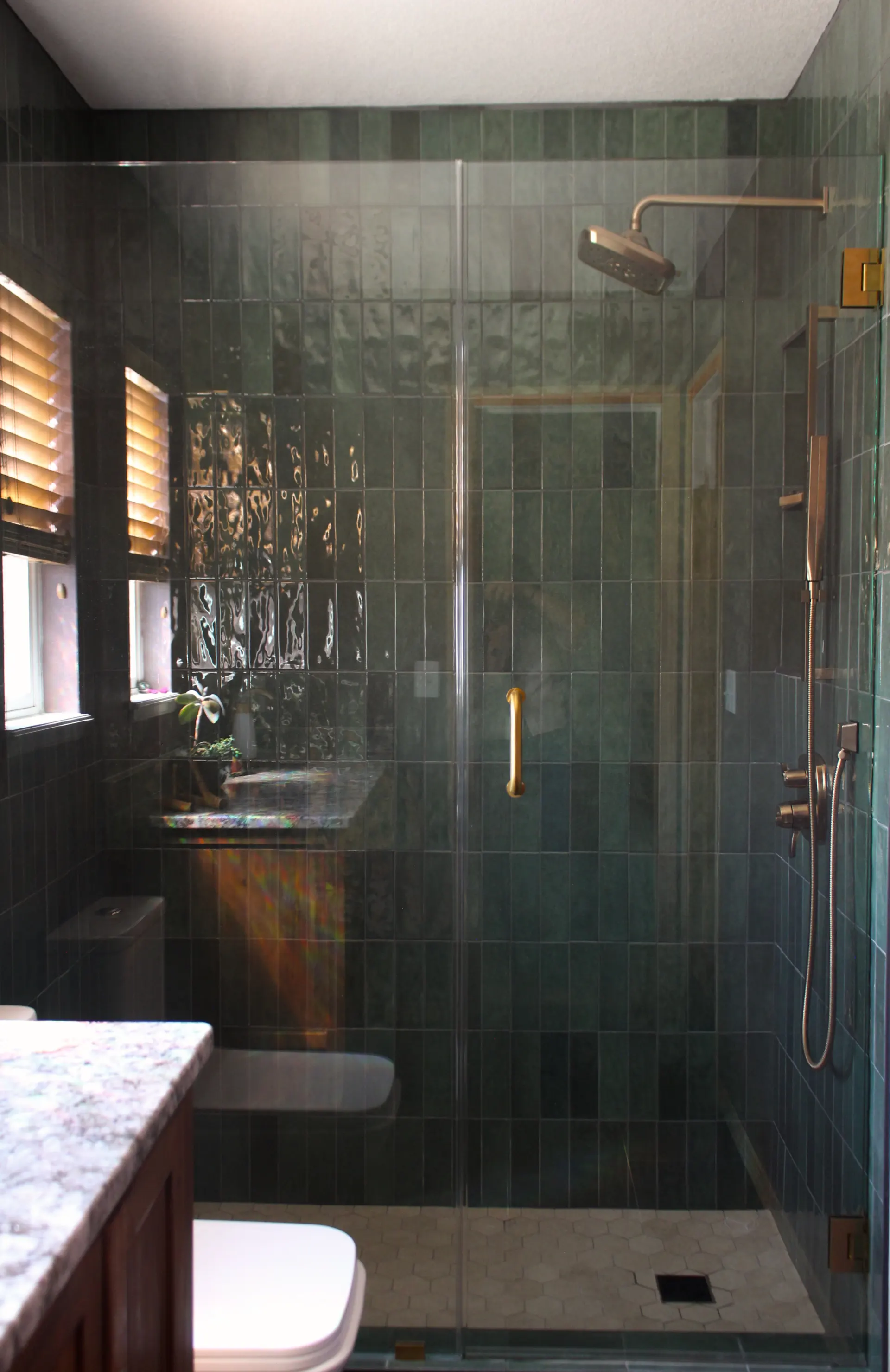 Bathroom with dark green tiled walls, a glass-enclosed shower with brass fixtures, and a white toilet next to a marble countertop.
