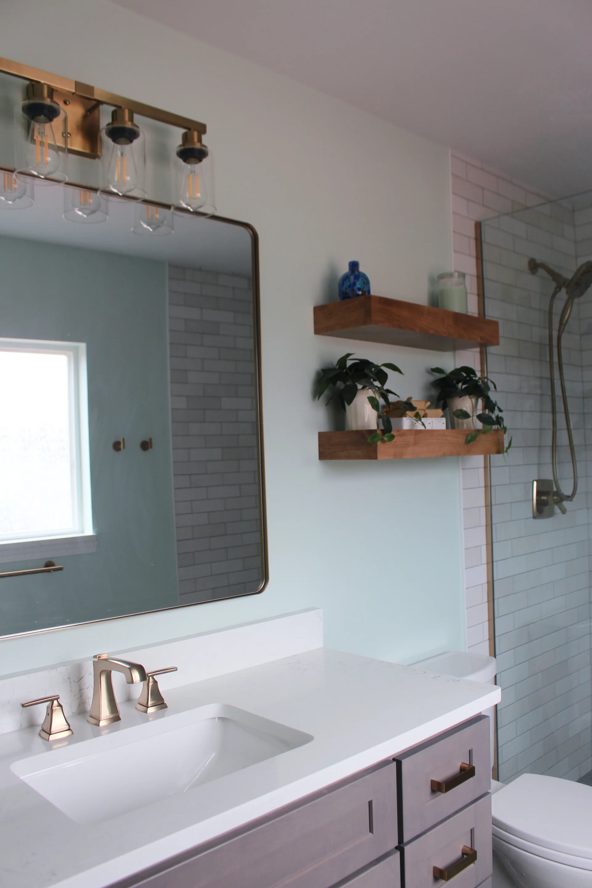 Bright bathroom with white countertop, gray wooden vanity, wall-mounted wooden shelves holding plants and decor, and glass-enclosed shower.