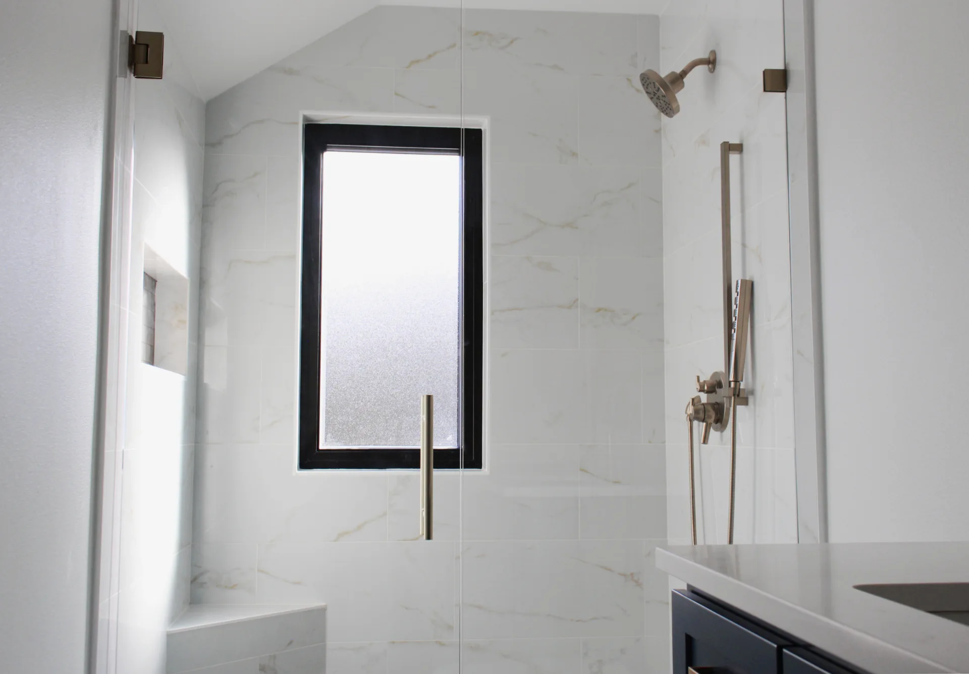 Modern bathroom shower with marble tiles, a frosted window, built-in bench, and brushed metal shower fixtures.