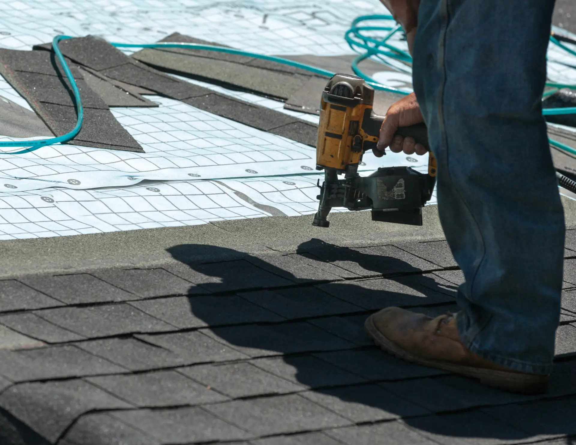 A worker in jeans and boots uses a nail gun to secure asphalt shingles on a roof. Loose shingles and a coiled hose are scattered nearby.