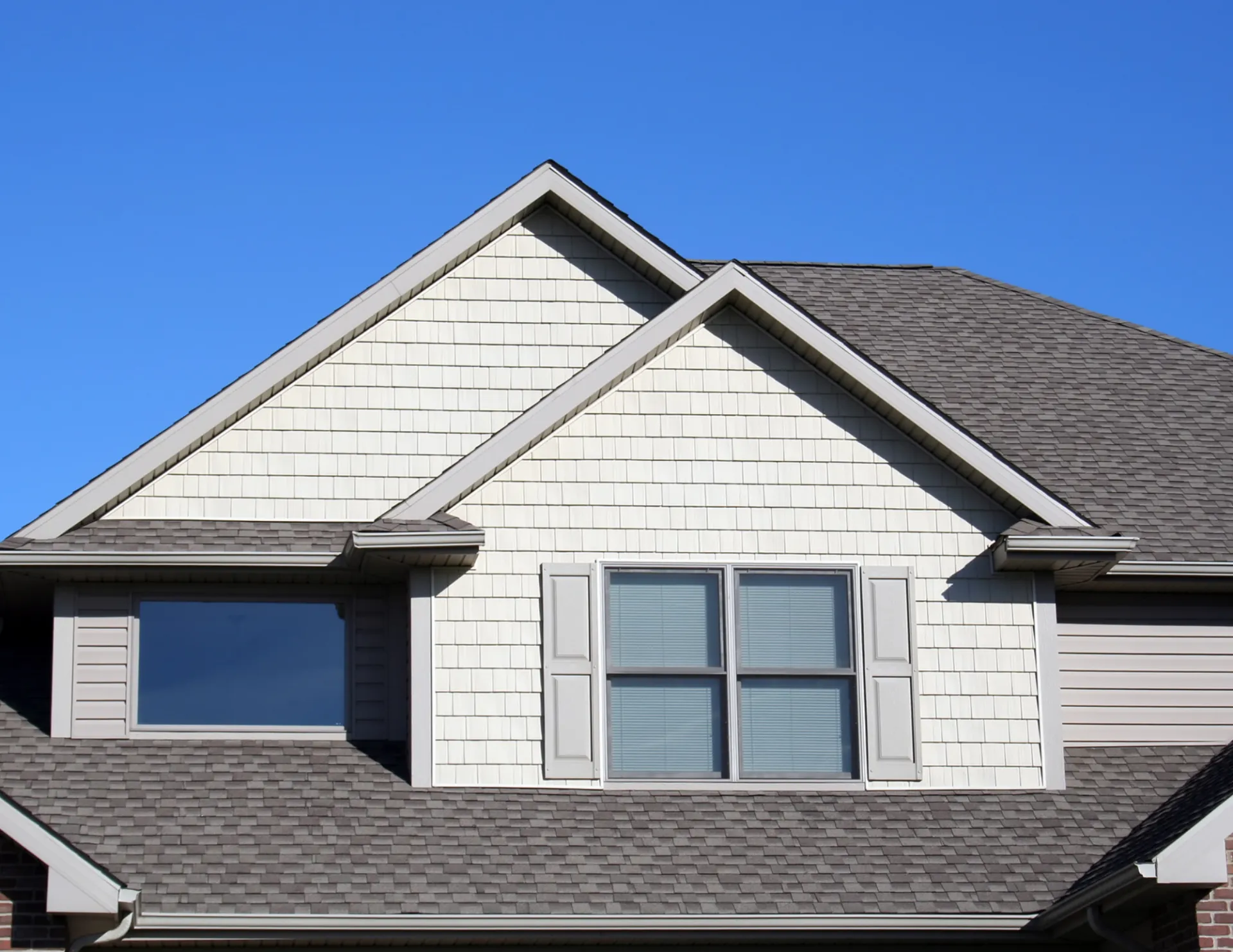 A suburban house rooftop with textured beige siding, two gabled peaks, shuttered windows, and a clear blue sky, conveying a calm and sunny day.