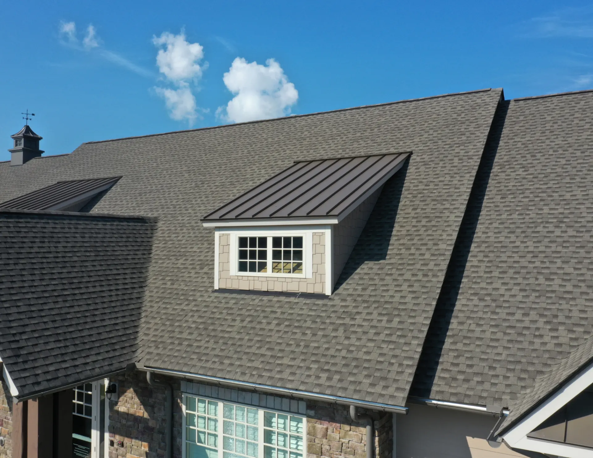 A close-up of a house roof with gray shingles and a metal-roofed dormer window under a clear blue sky. The scene is calm and well-maintained.