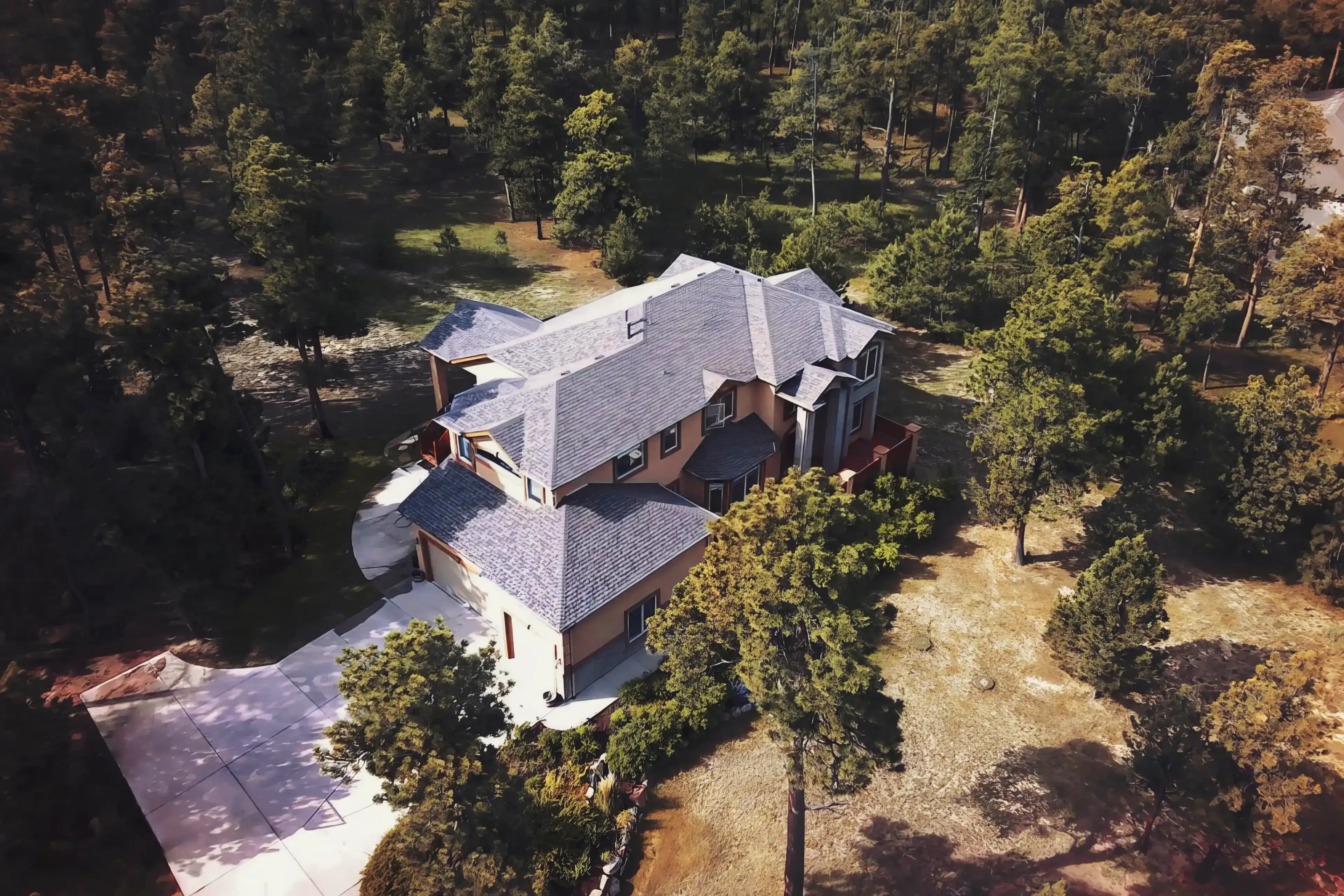 A large house with a gray shingle roof surrounded by trees and a concrete driveway in a wooded area.
