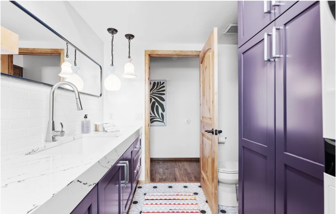 Modern bathroom with purple cabinets, white marble countertop, large mirror, and wooden door open to room with hardwood floor and wall art.