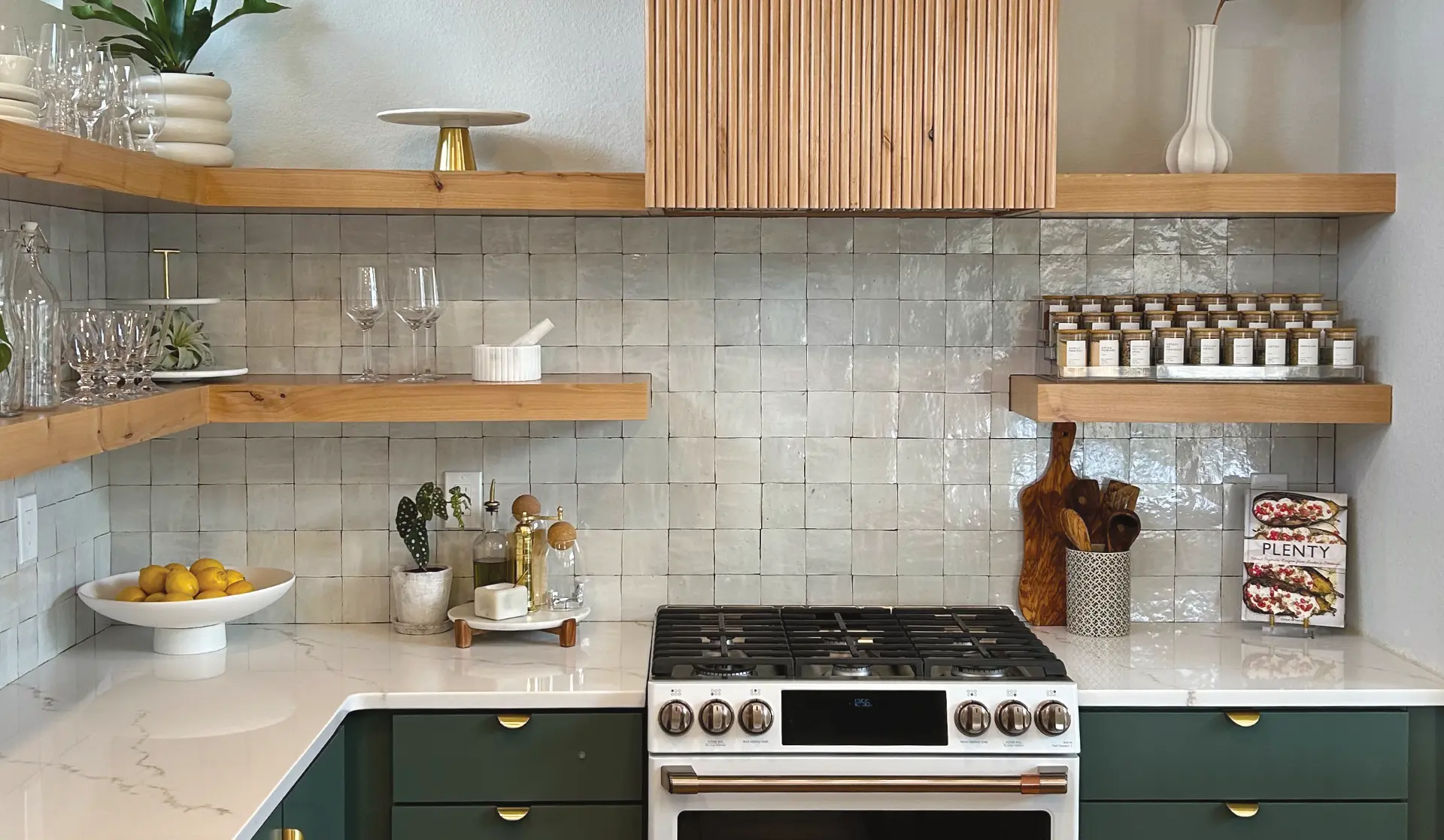 Modern kitchen with green lower cabinets, white marble countertops, wooden floating shelves holding glassware, spices, and kitchen utensils around a white gas stove with silver knobs.
