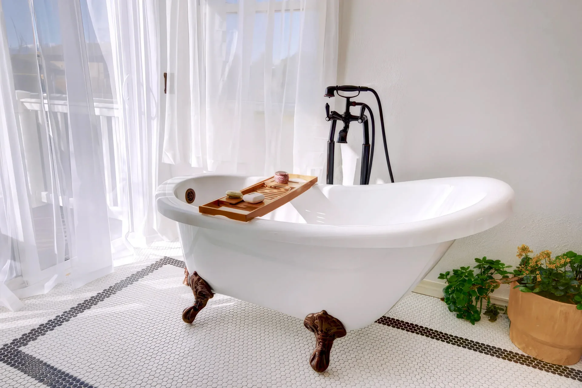 White freestanding clawfoot bathtub with dark bronze fixtures, a wooden bath tray with soap and candle, near sheer white curtains and a potted plant on mosaic tile floor.