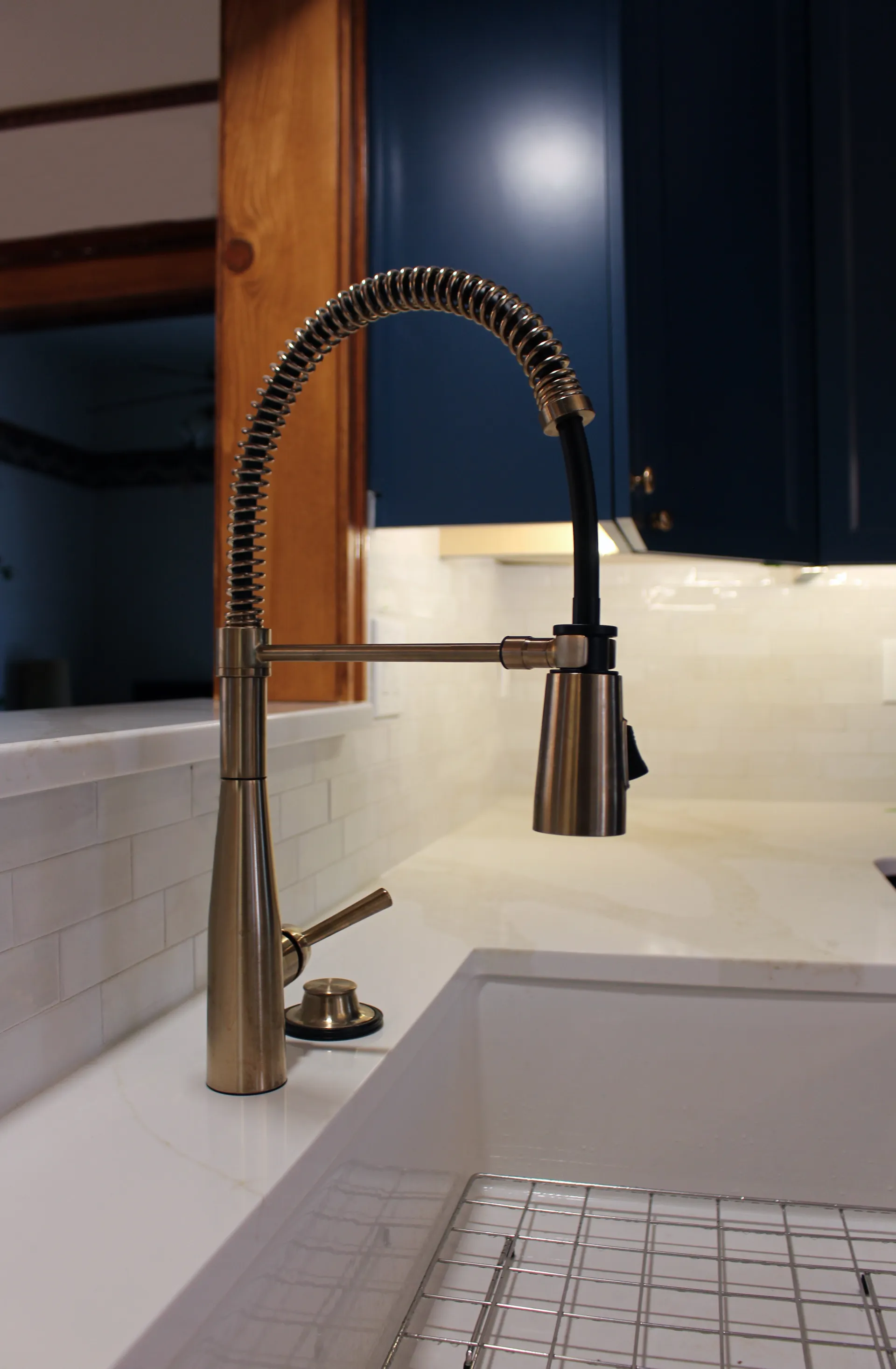 Modern kitchen sink faucet with flexible spring neck and gold finish mounted on white countertop with a white tile backsplash and blue cabinets in the background.