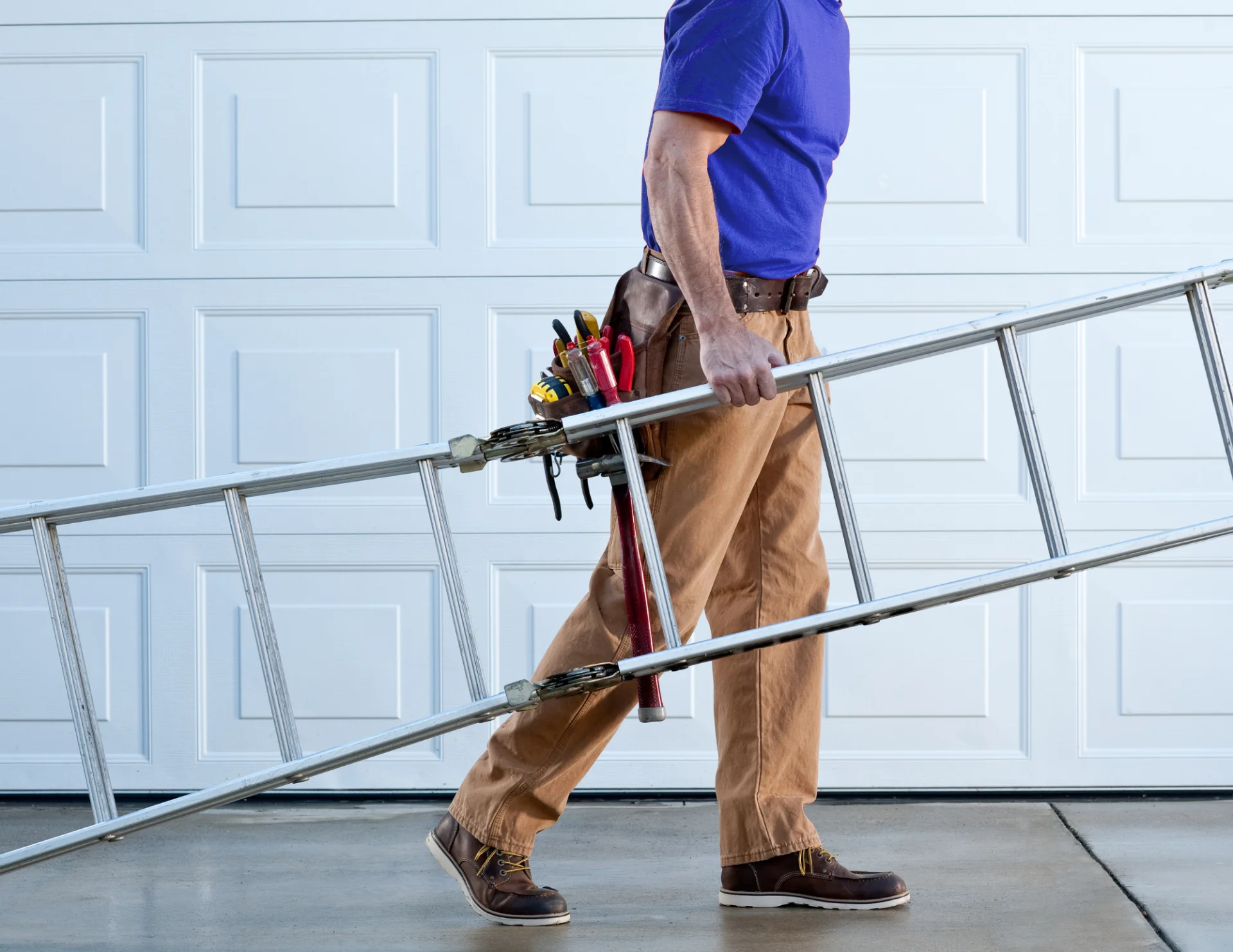 Man wearing brown pants, blue shirt, and tool belt carrying an aluminum ladder in front of a garage door.