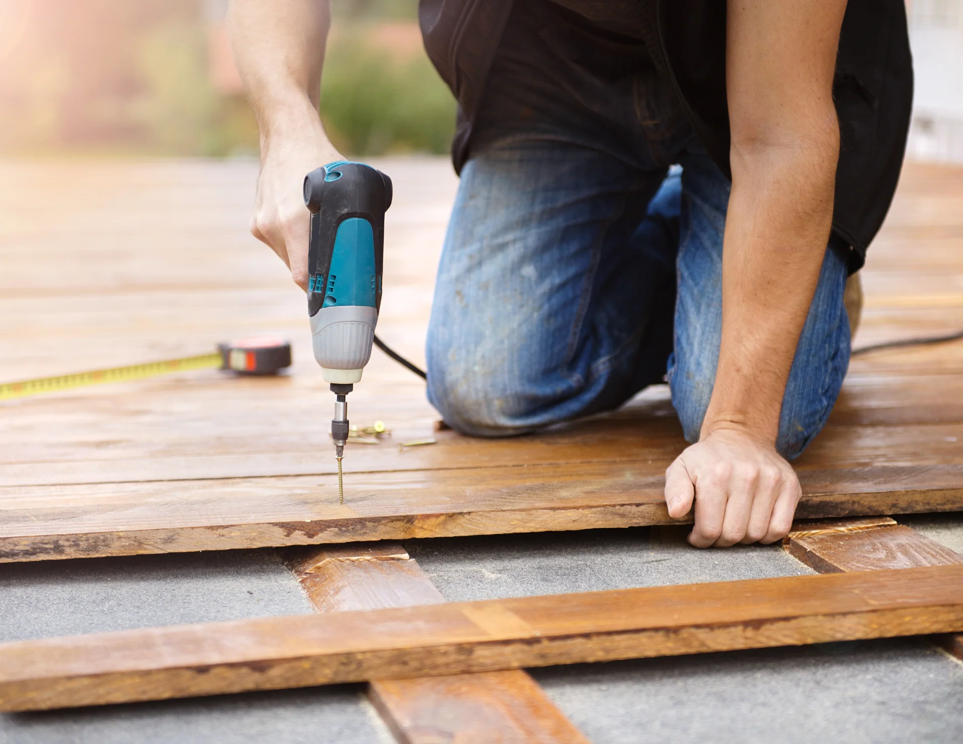 Person kneeling on wooden decking using a power drill to screw down a plank.