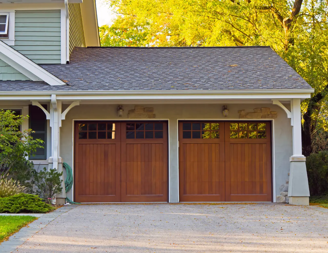 Modern house with two wooden garage doors under a gray shingled roof surrounded by greenery.