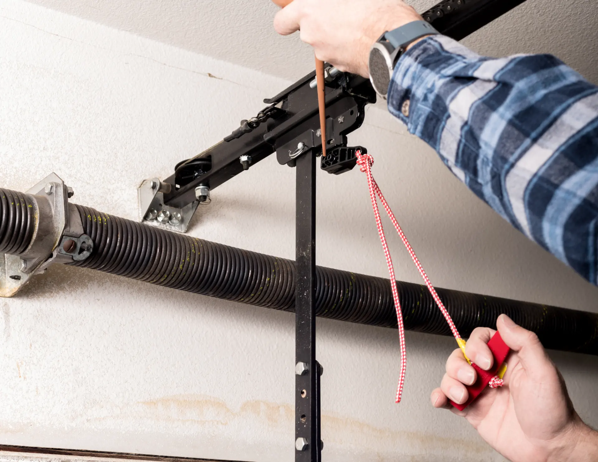 Person's hands repairing a garage door mechanism, holding a red pull cord and using a screwdriver on the black metal track.