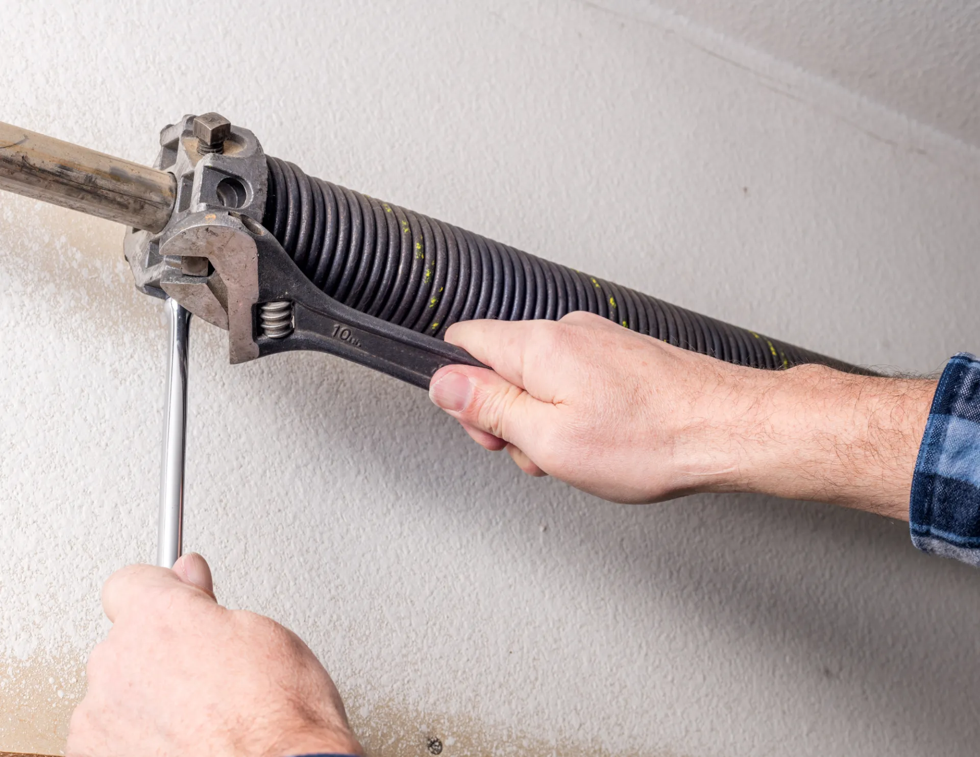 Hands using a wrench and screwdriver to adjust a black garage door torsion spring mounted on a wall.