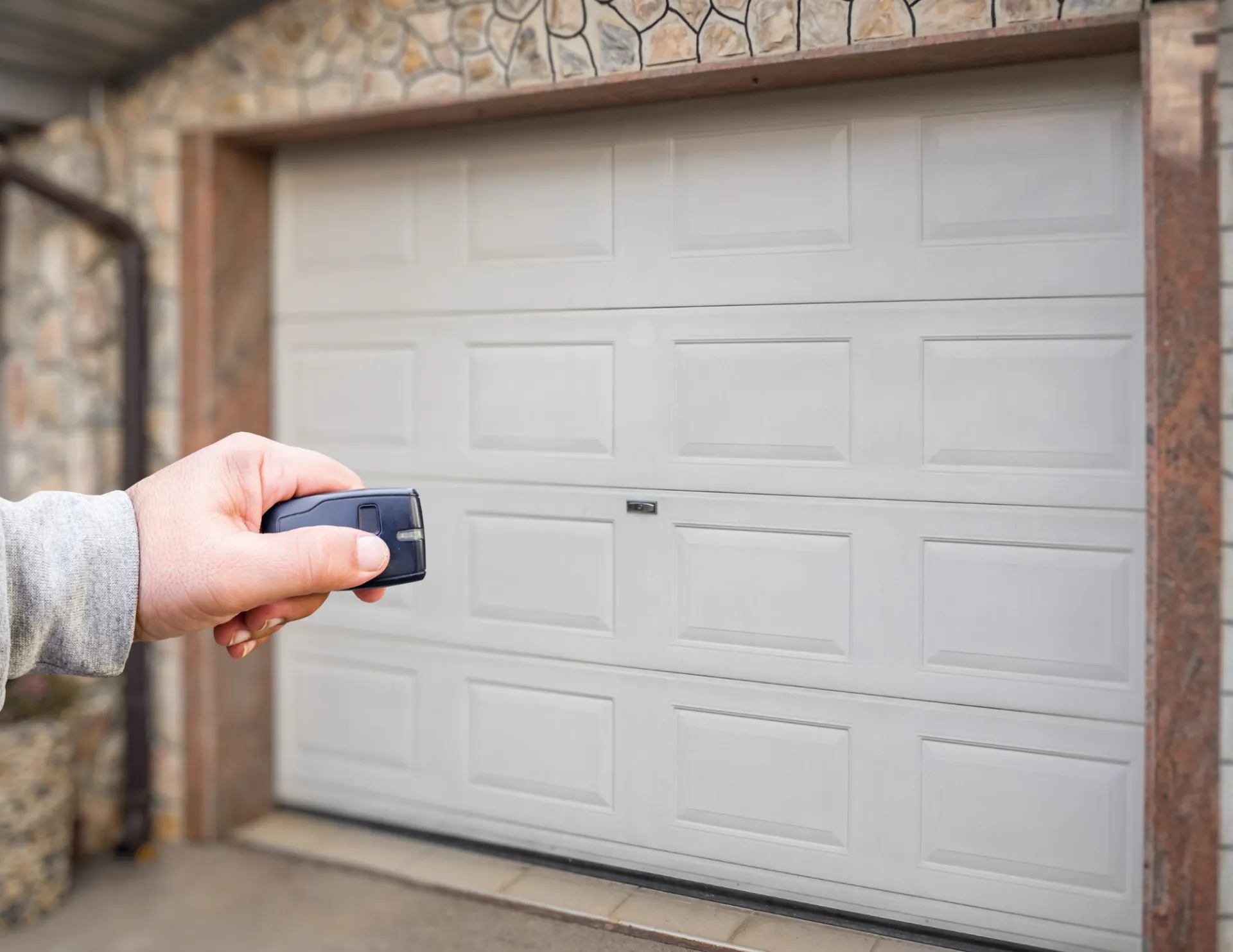 Person holding a remote control aimed at a closed white garage door with a stone exterior.