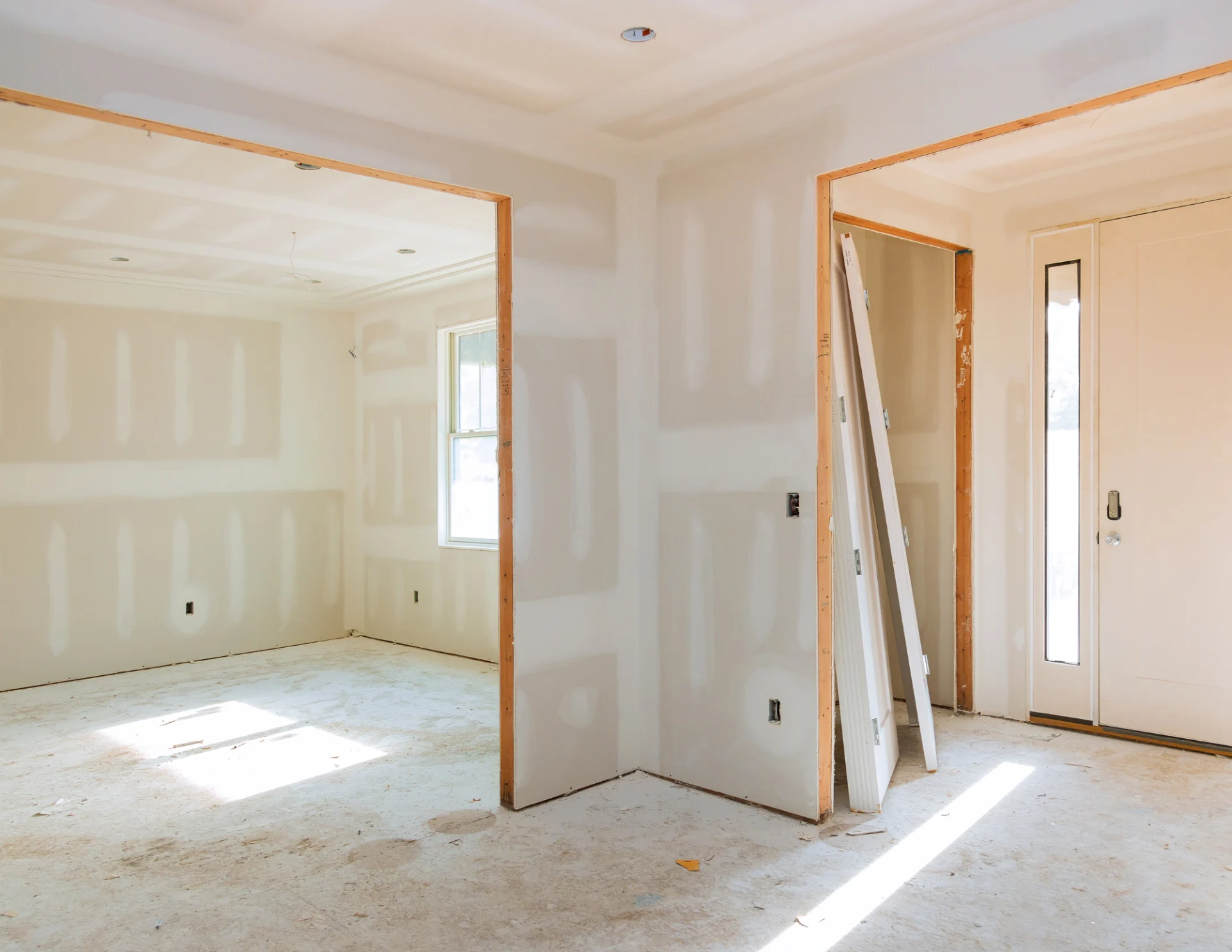 Interior of an unfinished room with drywall installed and unpainted, with sunlight streaming through a window and an inbox door leaning against a wall.