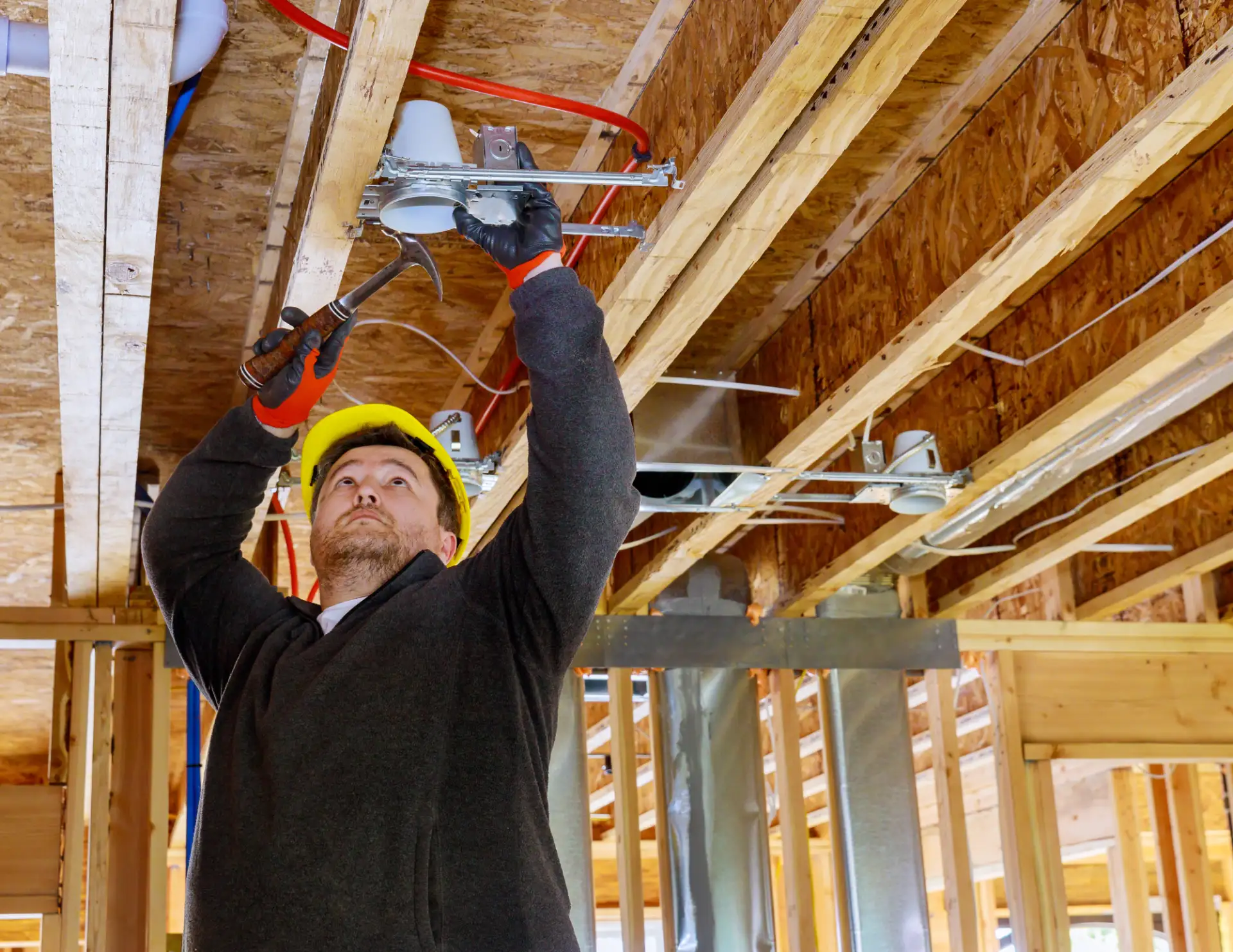 Electrician wearing a yellow hard hat and gloves installing ceiling electrical fixtures in a building under construction.