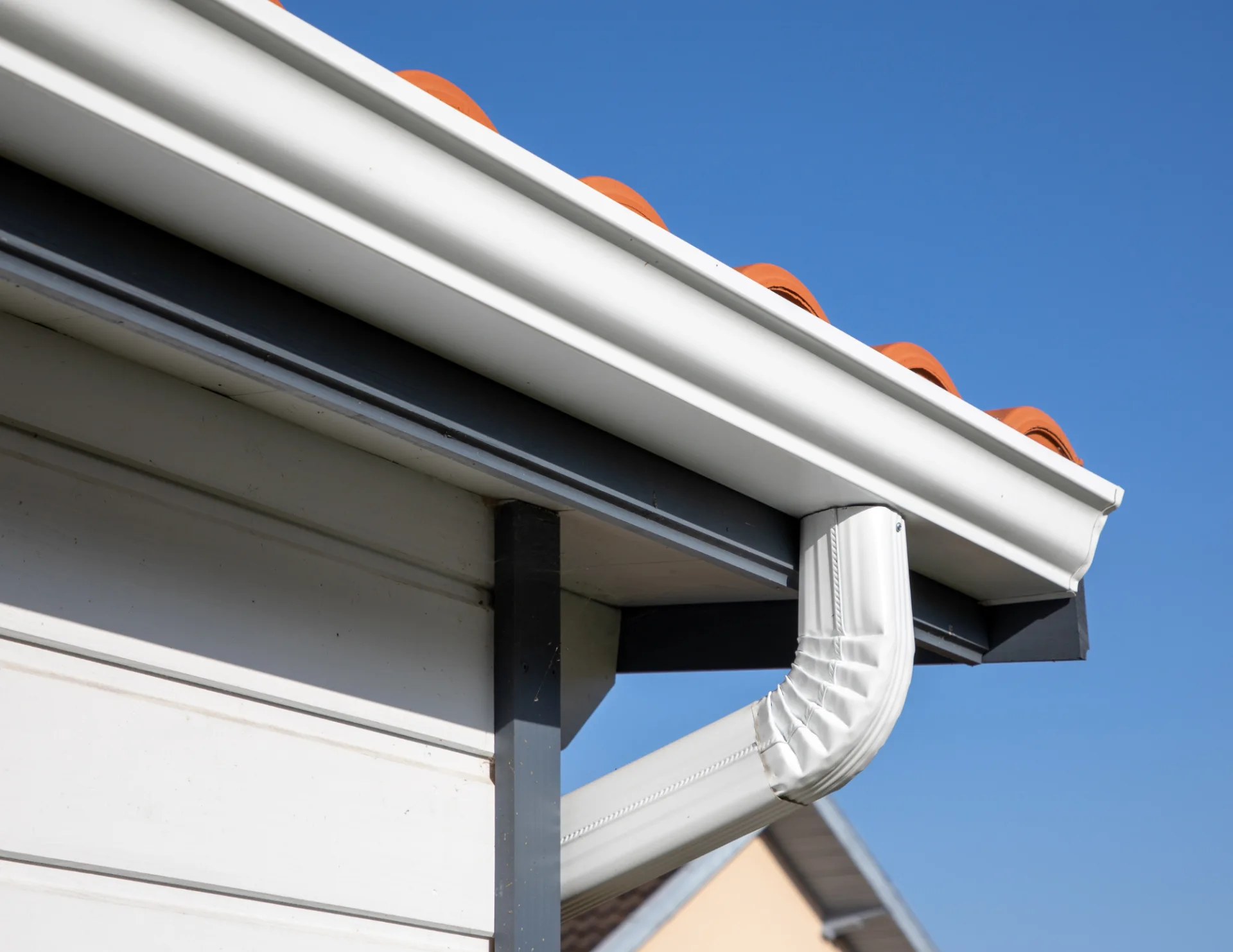 Close-up of a white gutter and downspout on a house with terracotta roof tiles against a clear blue sky.