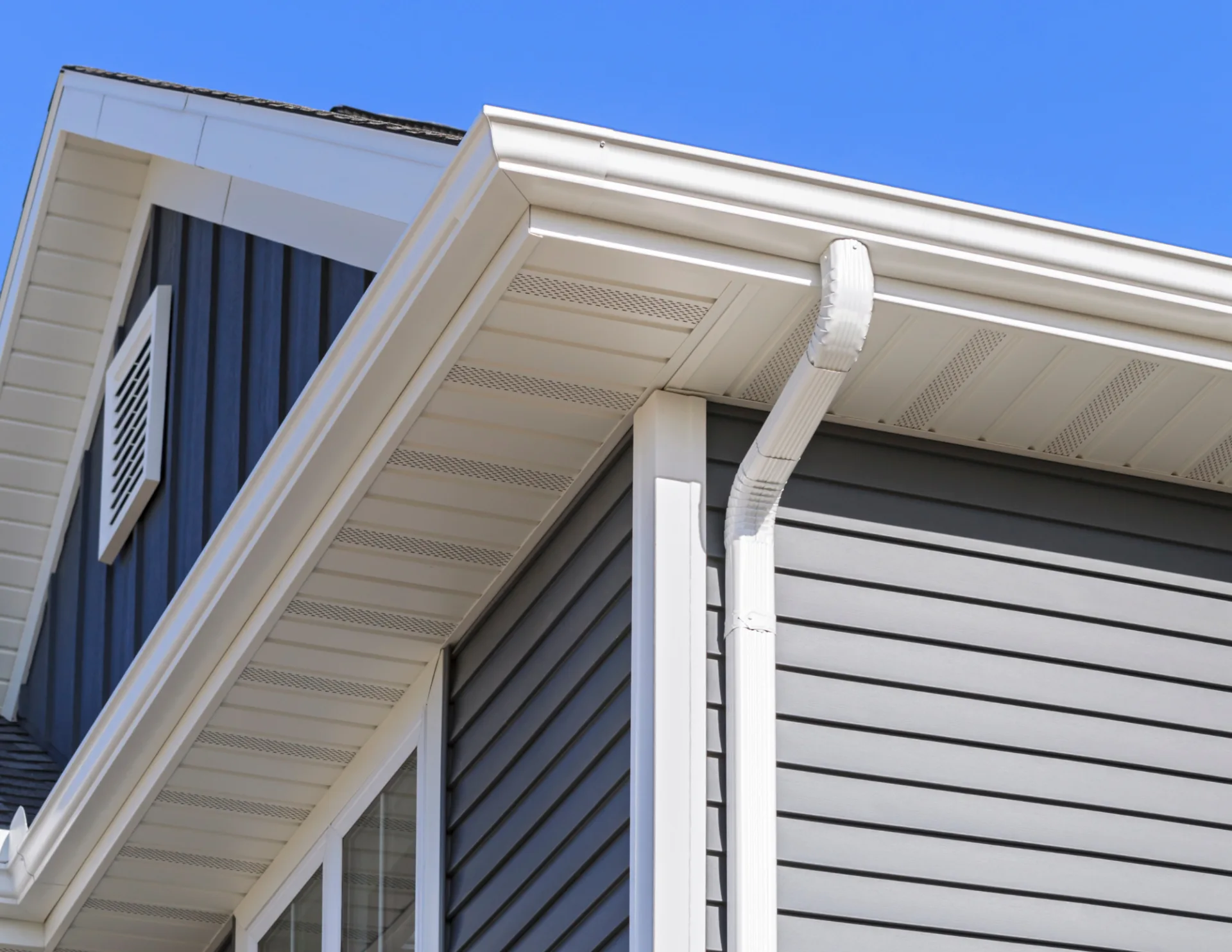 Close-up of house corner showing white gutters and downspouts above gray horizontal siding under a clear blue sky.