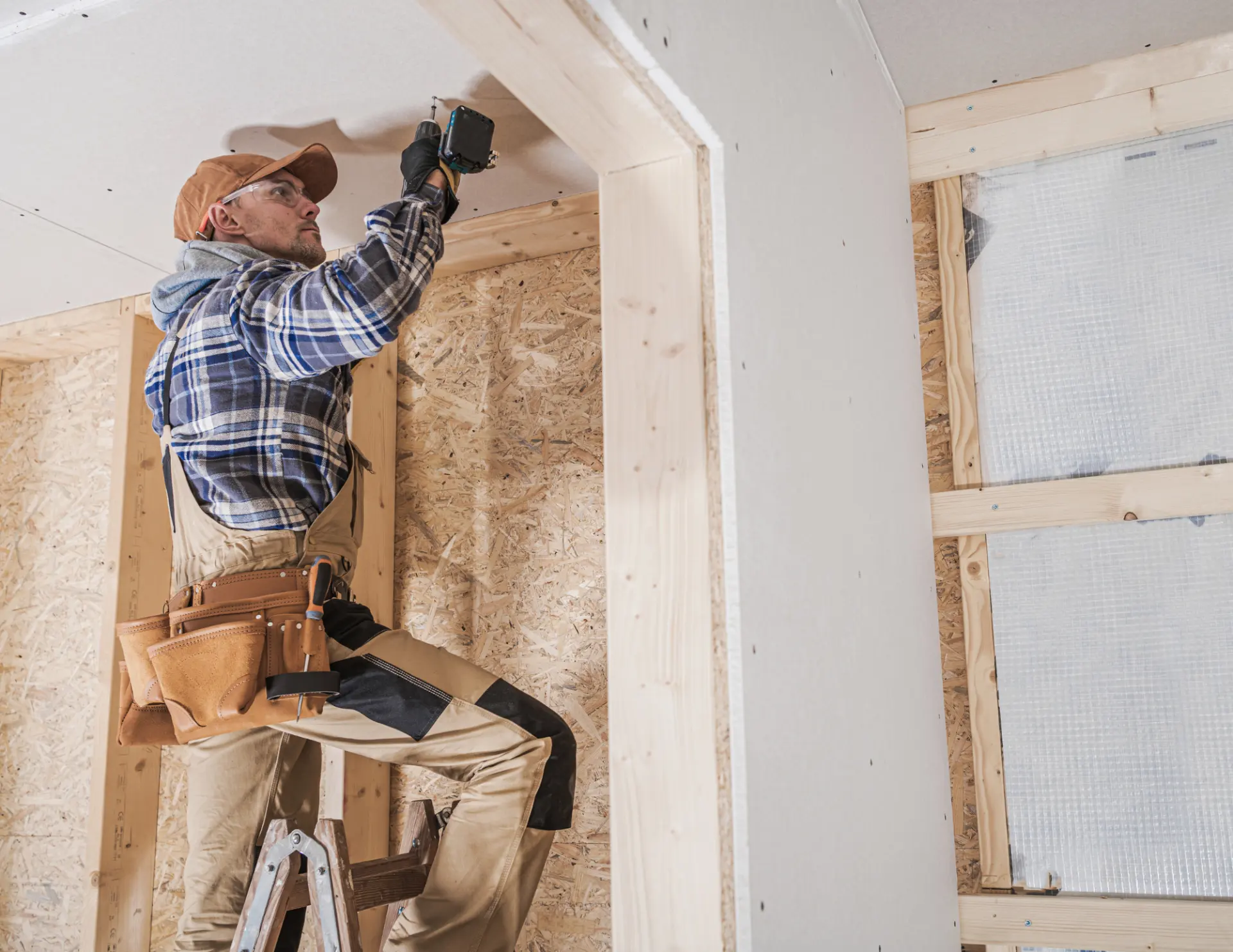 Construction worker in a plaid shirt and cap standing on a ladder, installing drywall inside a wooden frame structure.