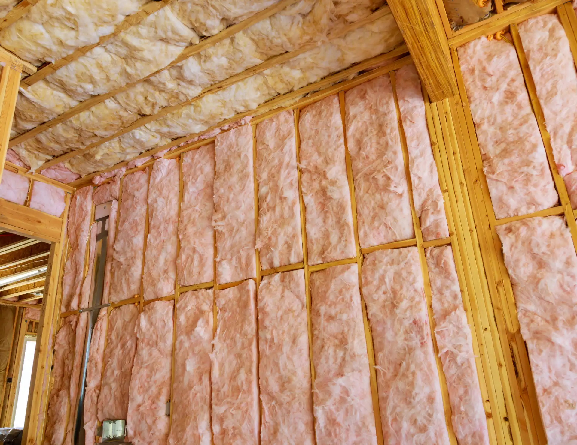 Interior wooden wall and ceiling studs filled with pink and yellow fiberglass insulation batts.