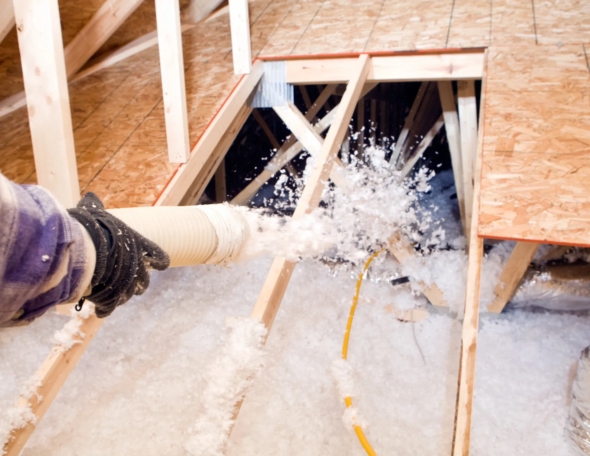 Hand spraying loose-fill insulation into an attic with exposed wooden beams.