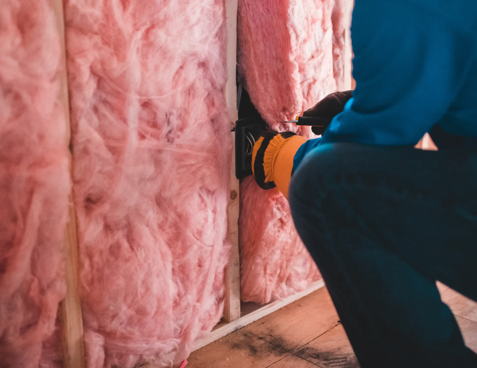 Person wearing blue and yellow gloves installing pink fiberglass insulation around an electrical outlet in a wall.